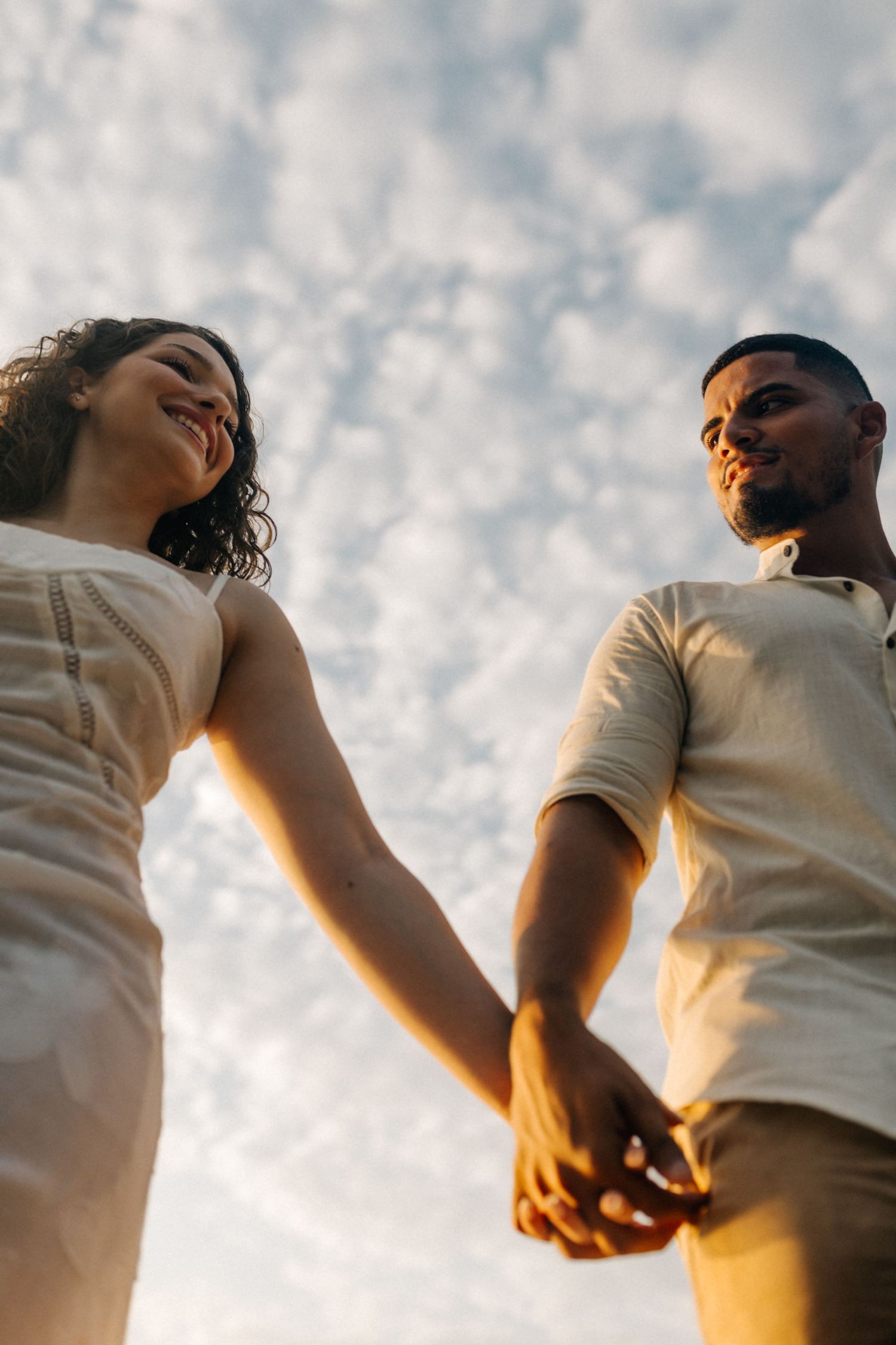 A couple in white outfits holds hands against a cloudy sky backdrop at sunset.