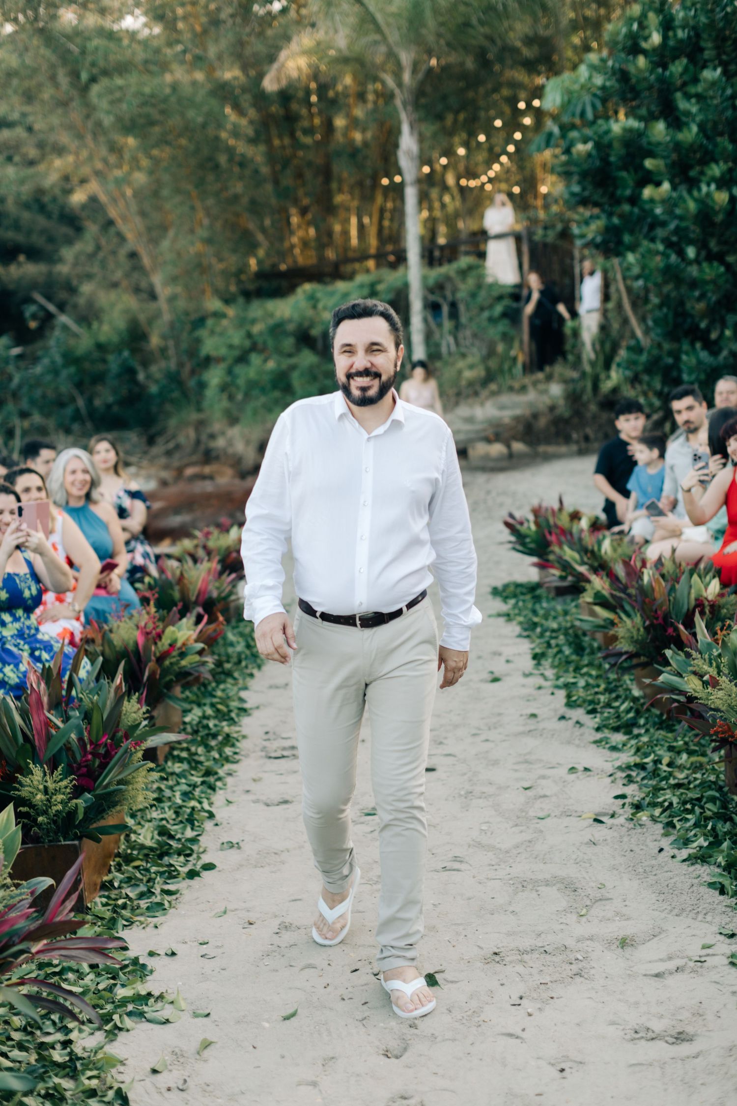 Person in white shirt and grey pants walking down a sandy aisle at outdoor ceremony.