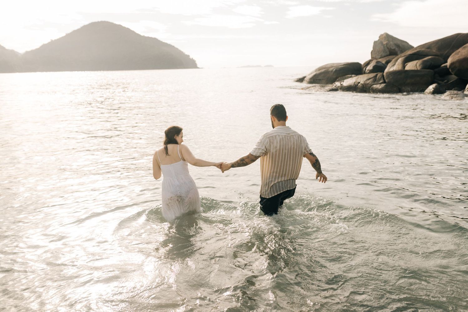 A couple wades through ocean water at a scenic beach during sunset.