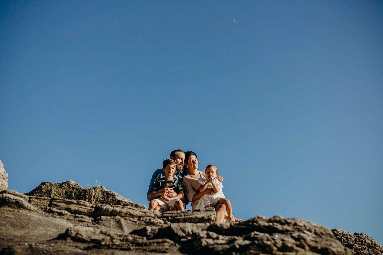 Family lounging on rocky coastline against blue sky and ocean backdrop.