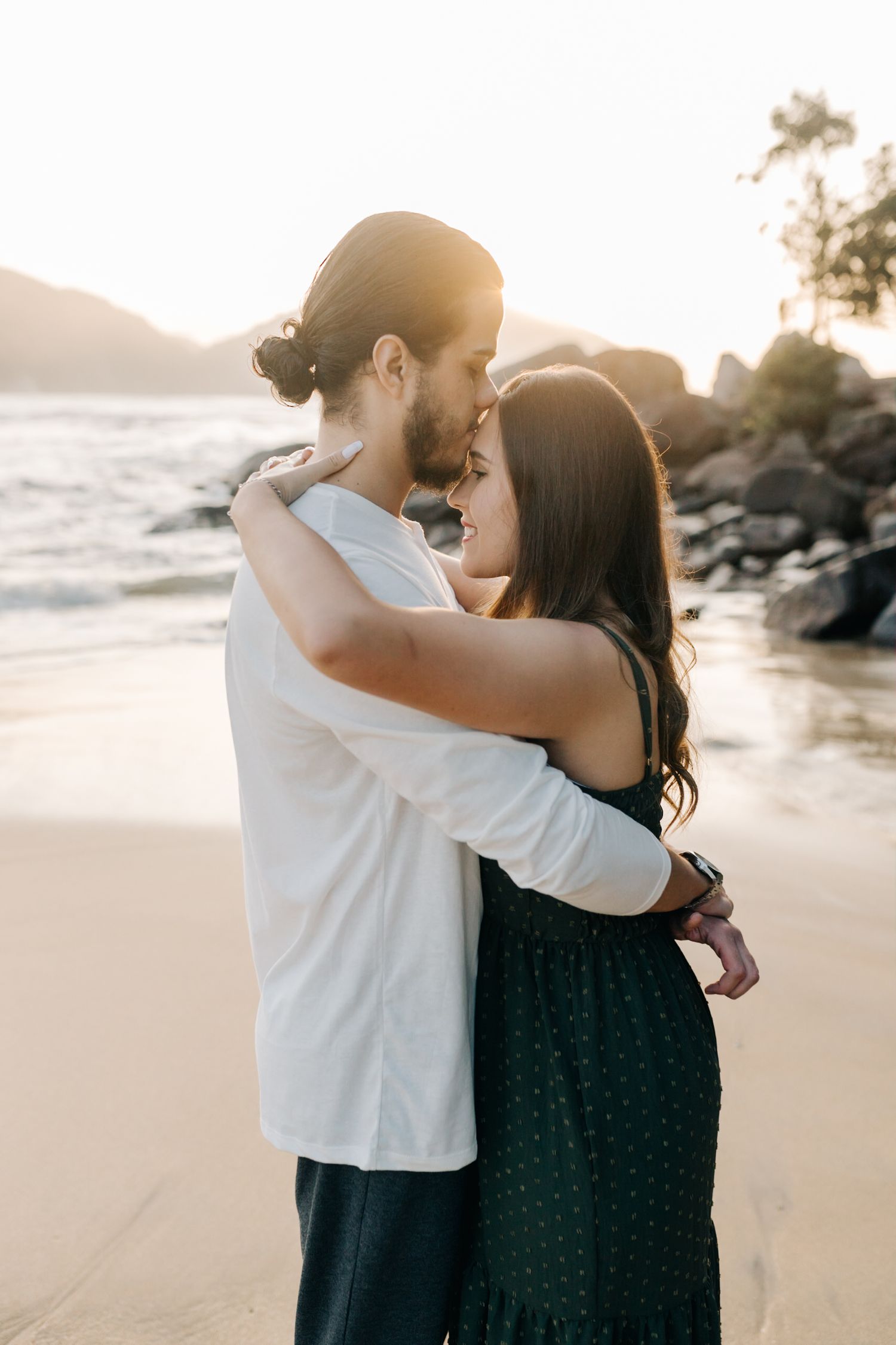 Intimate moment between couple sharing embrace during golden hour on secluded beach with rocky backdrop.