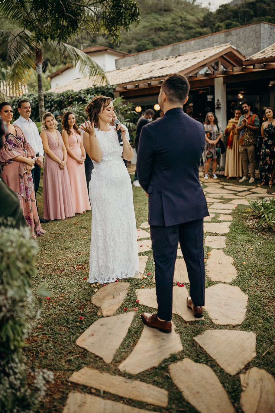 A bride and groom stand on stone pathway during outdoor wedding ceremony with bridesmaids in pink dresses.
