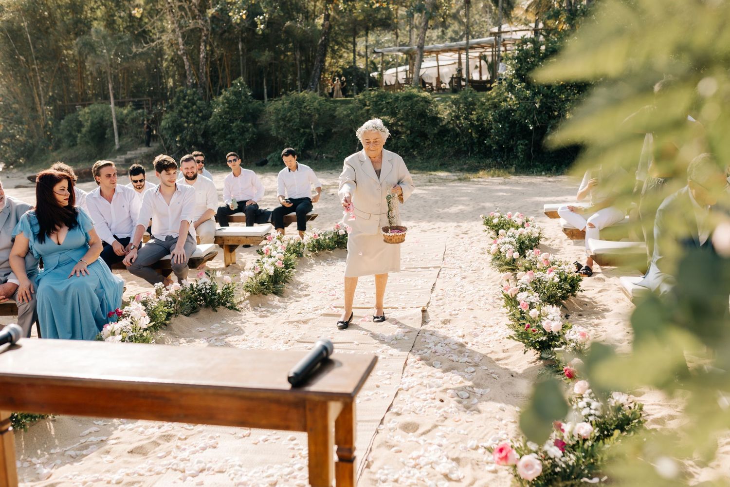 An outdoor wedding ceremony with guests seated on benches surrounded by pink floral arrangements.