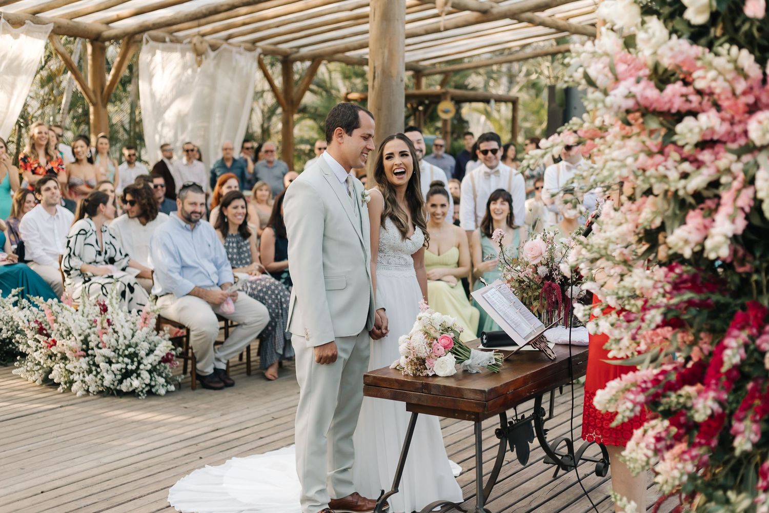 Wedding celebration under wooden beams with pink flower arrangements and gathered guests.