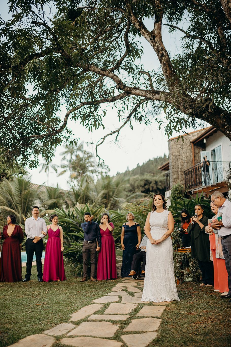 Wedding ceremony outdoors with attendees in colorful dresses standing along a stone path near a rustic building.