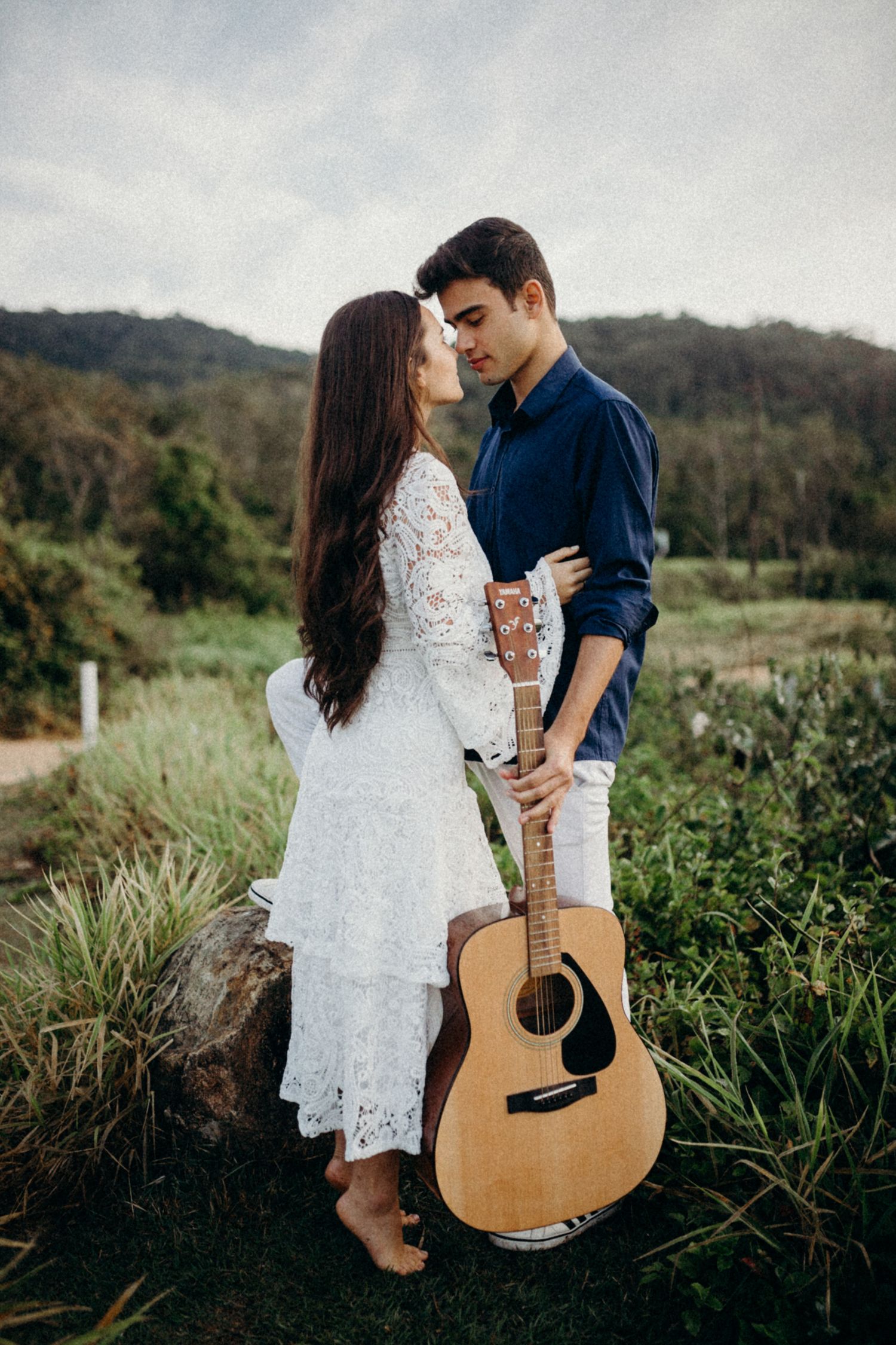 A romantic couple pose with an acoustic guitar against a scenic mountain backdrop.