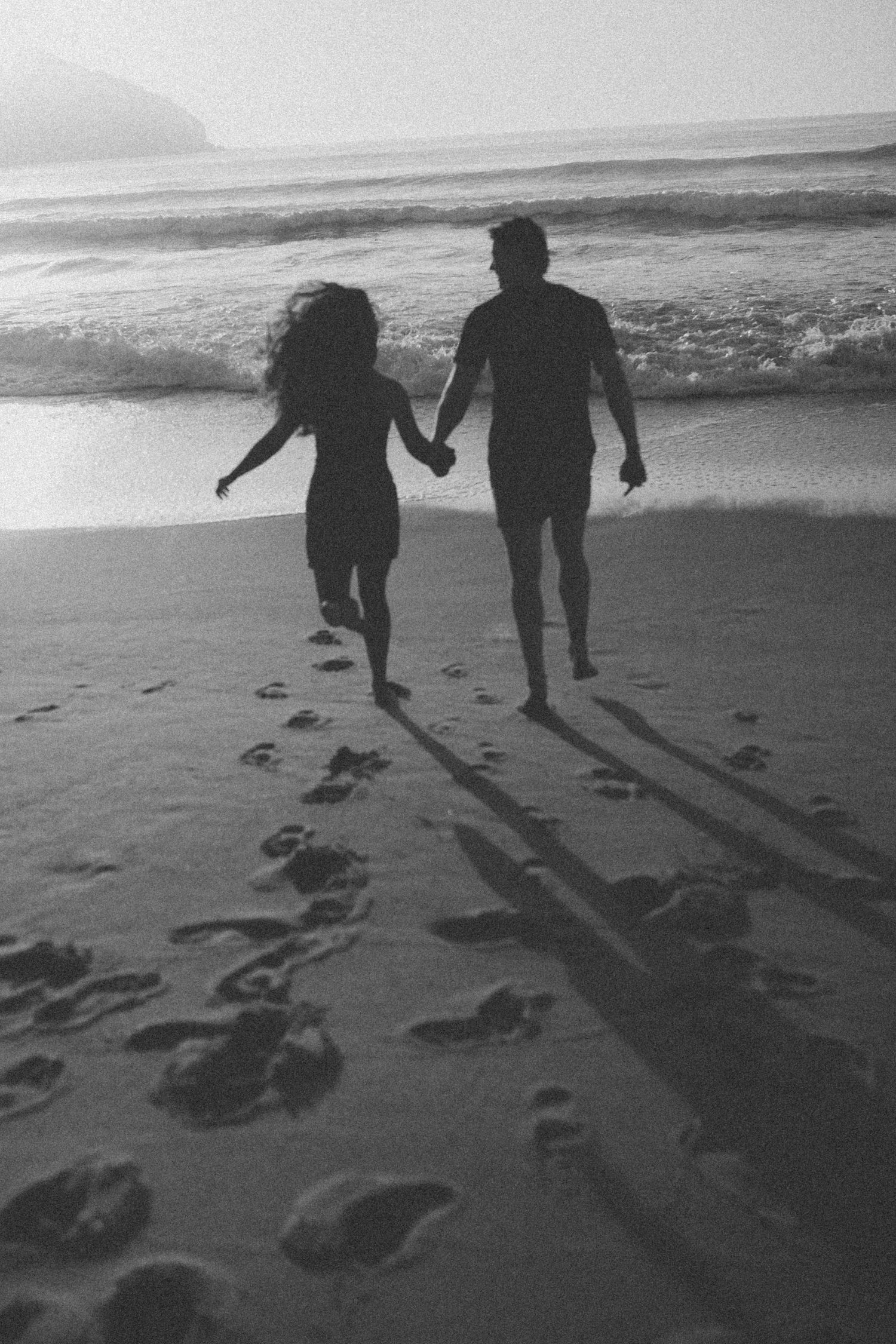 Black and white photo of couple walking on beach leaving footprints in sand.