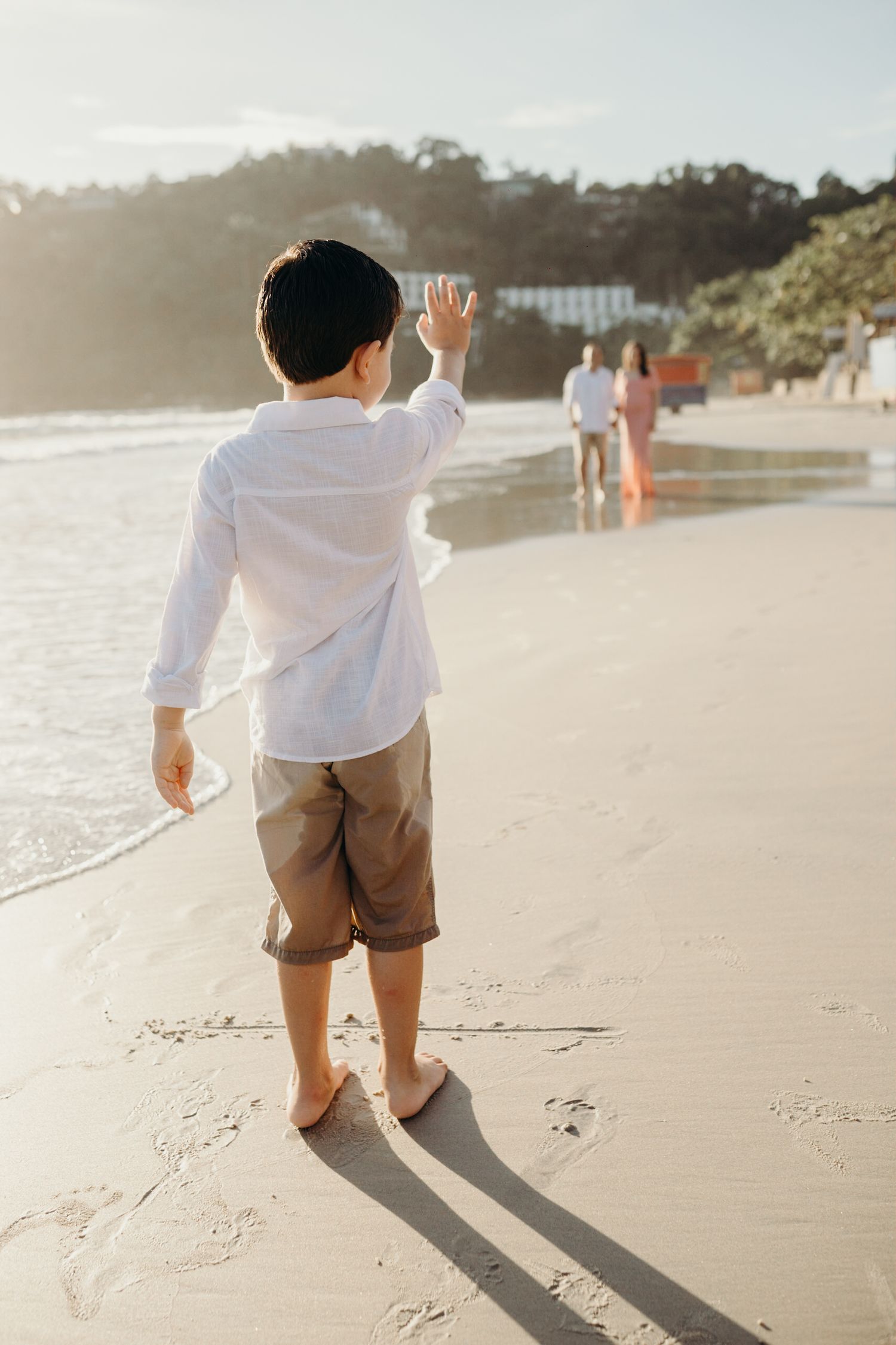 Child in white shirt waves from beach as others walk in distance during sunset.