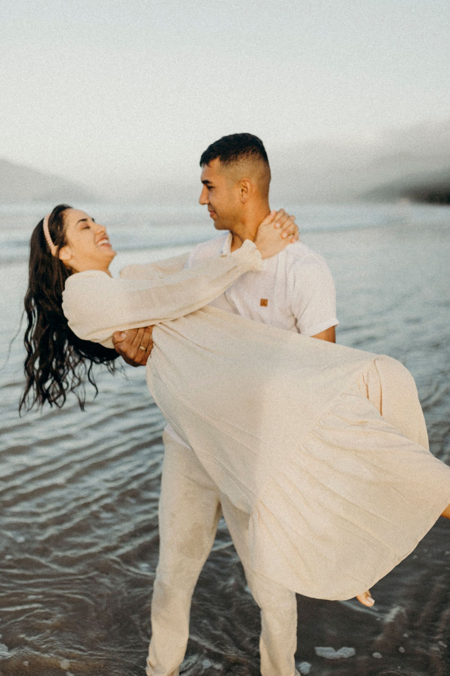 A couple dressed in white embraces while standing in shallow ocean water during golden hour.