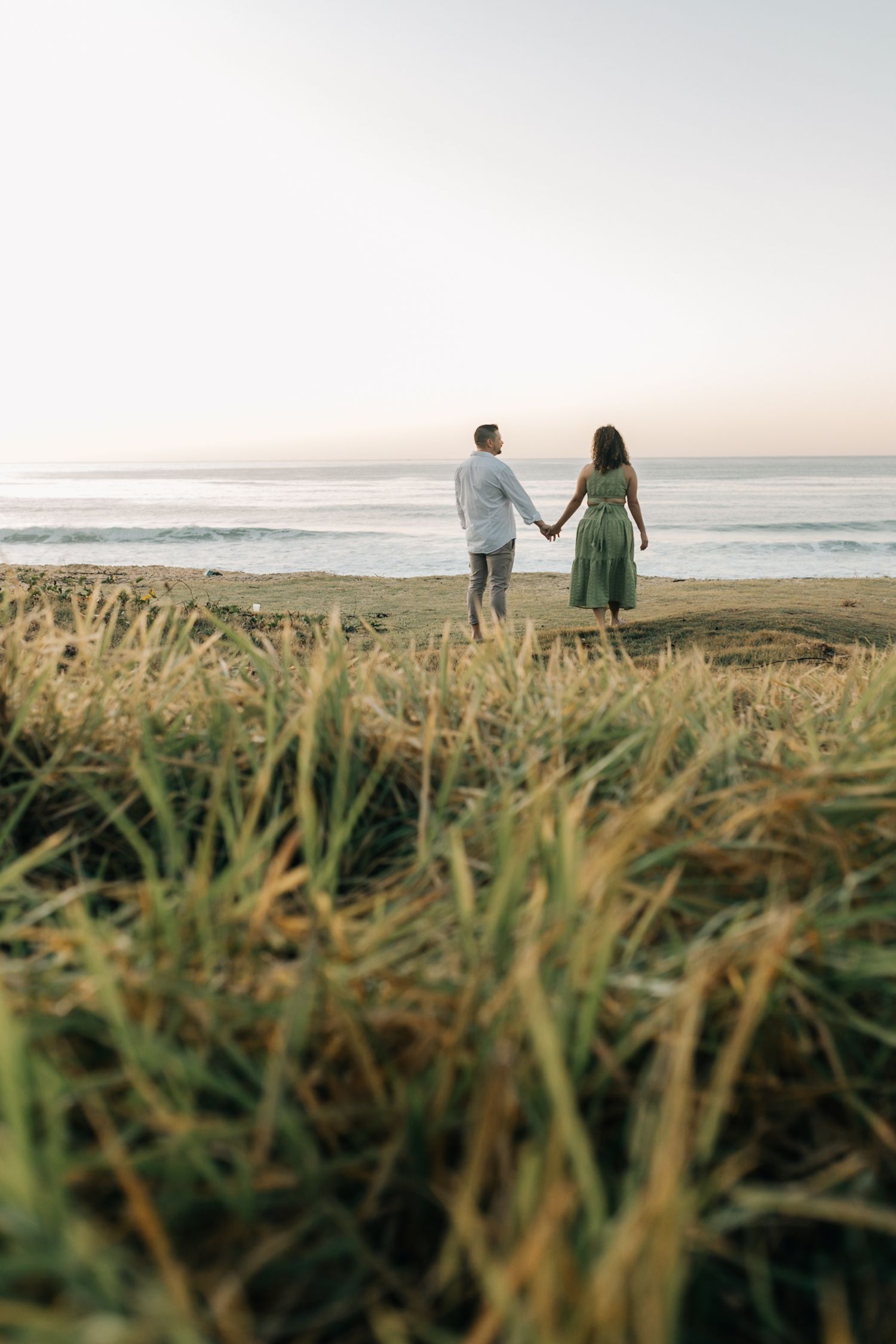 Couple walking hand in hand through tall grass overlooking ocean at sunset.