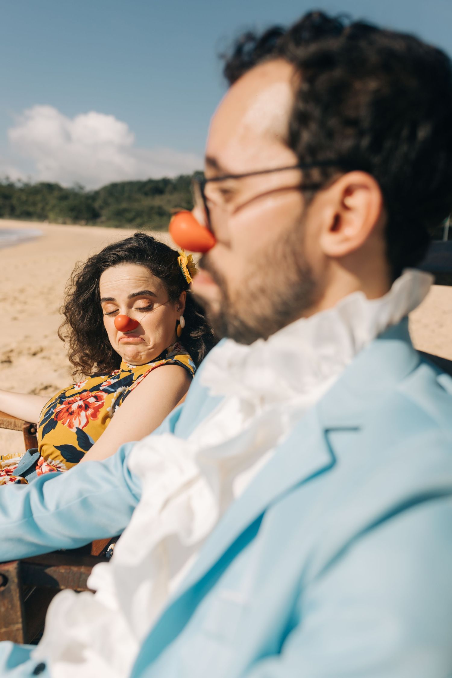 A couple wearing red noses shares a playful moment in beach chairs.