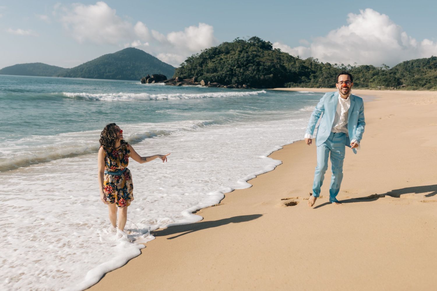 A couple plays in the ocean waves on a scenic beach with mountains in the background.