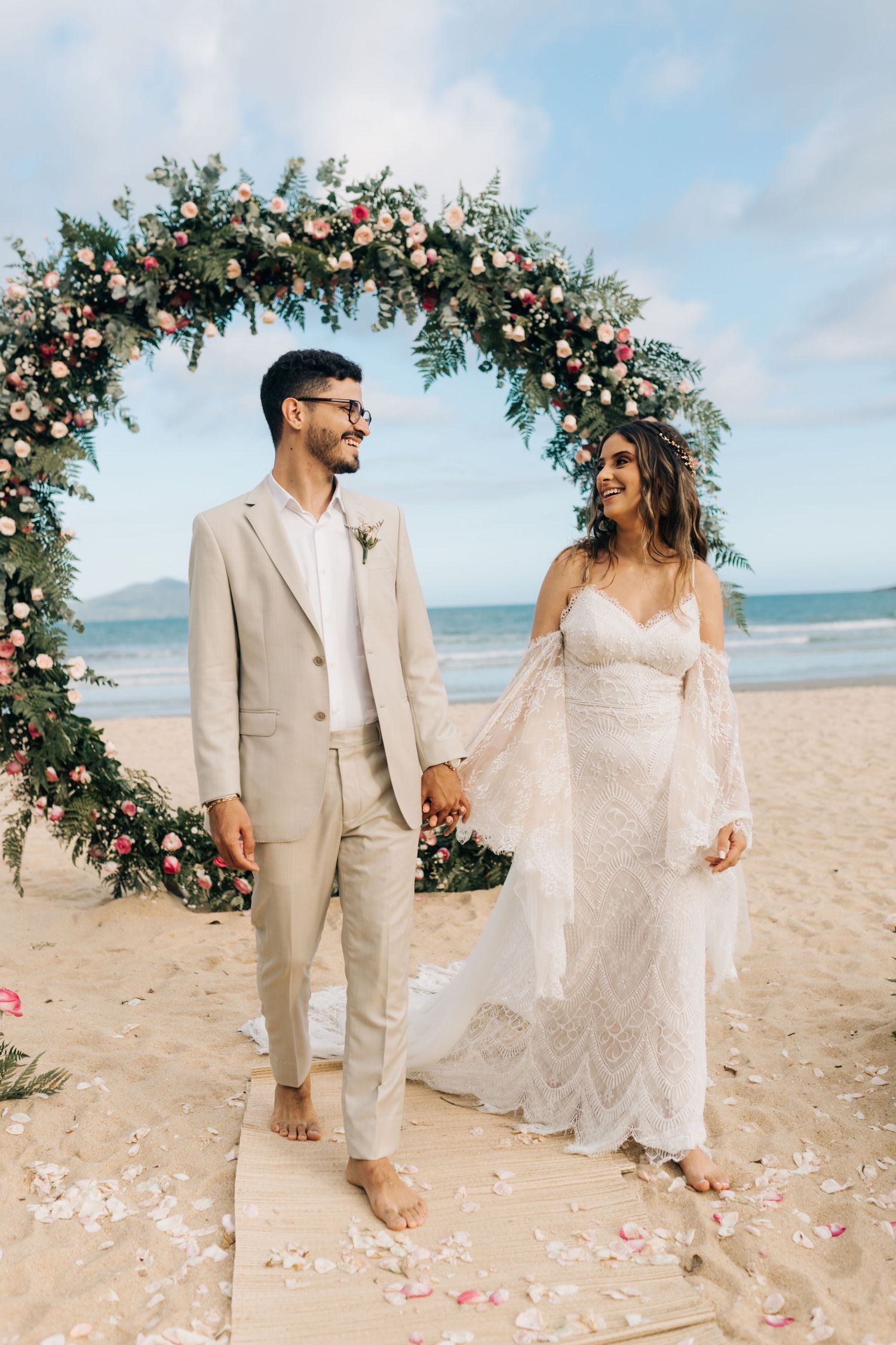 Newlyweds pose under floral arch during beachside ceremony with ocean view.