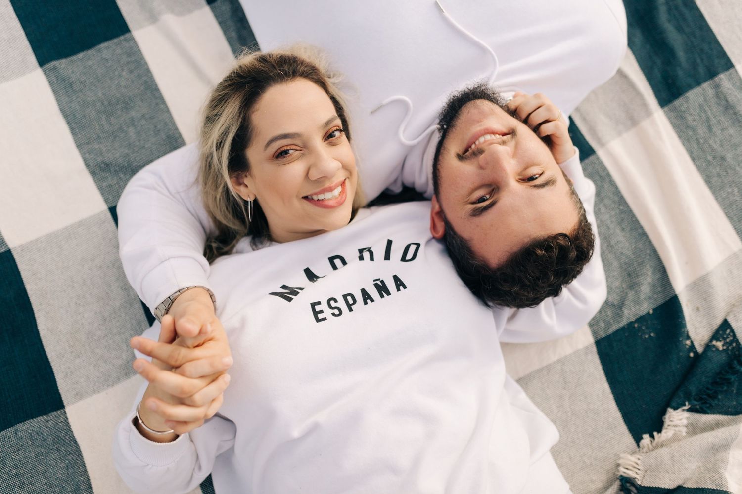 Happy couple holding hands while lying on checkered blanket wearing matching shirts.