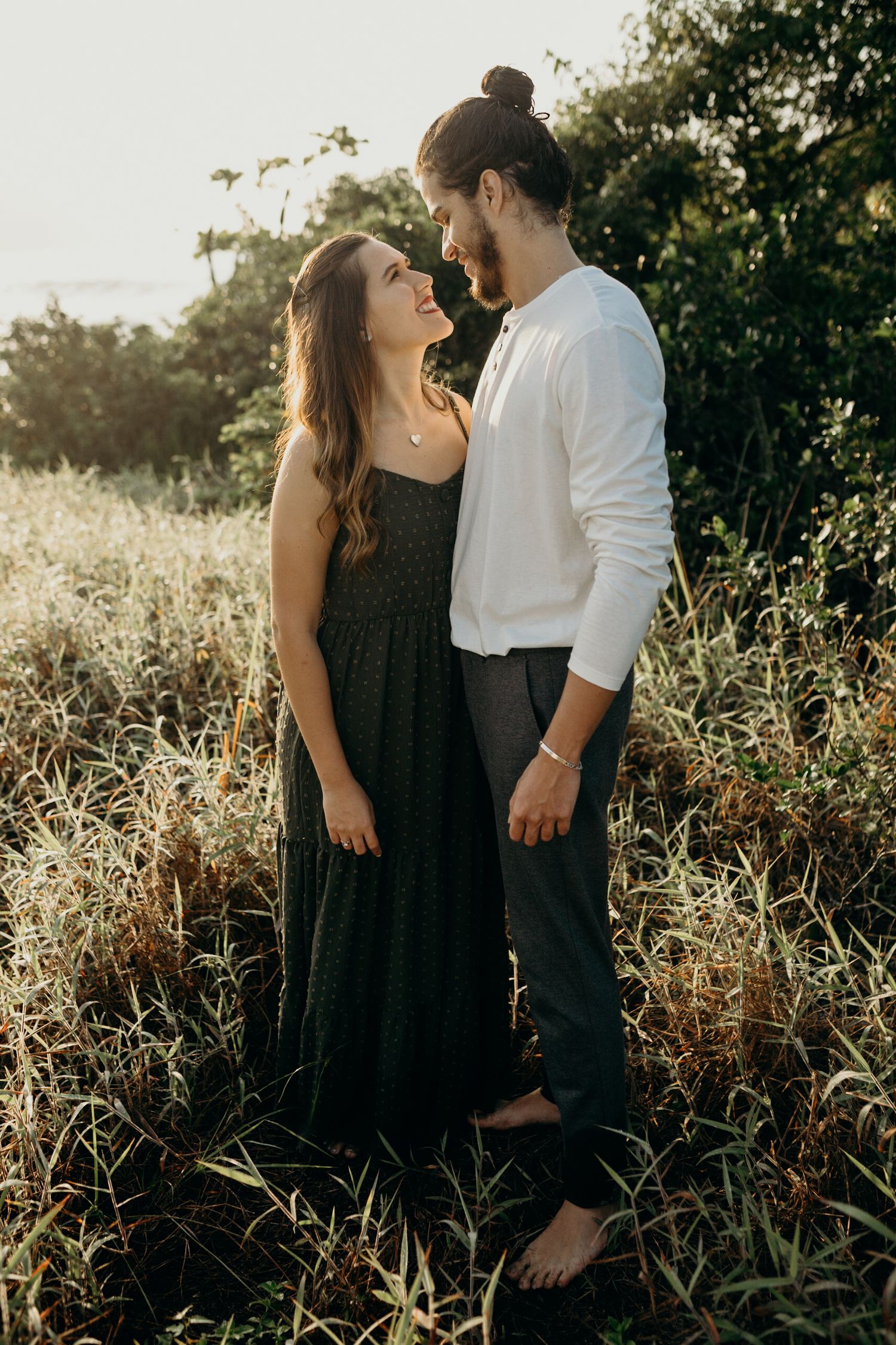 Romantic couple in black dress and white shirt poses barefoot in sunlit meadow.
