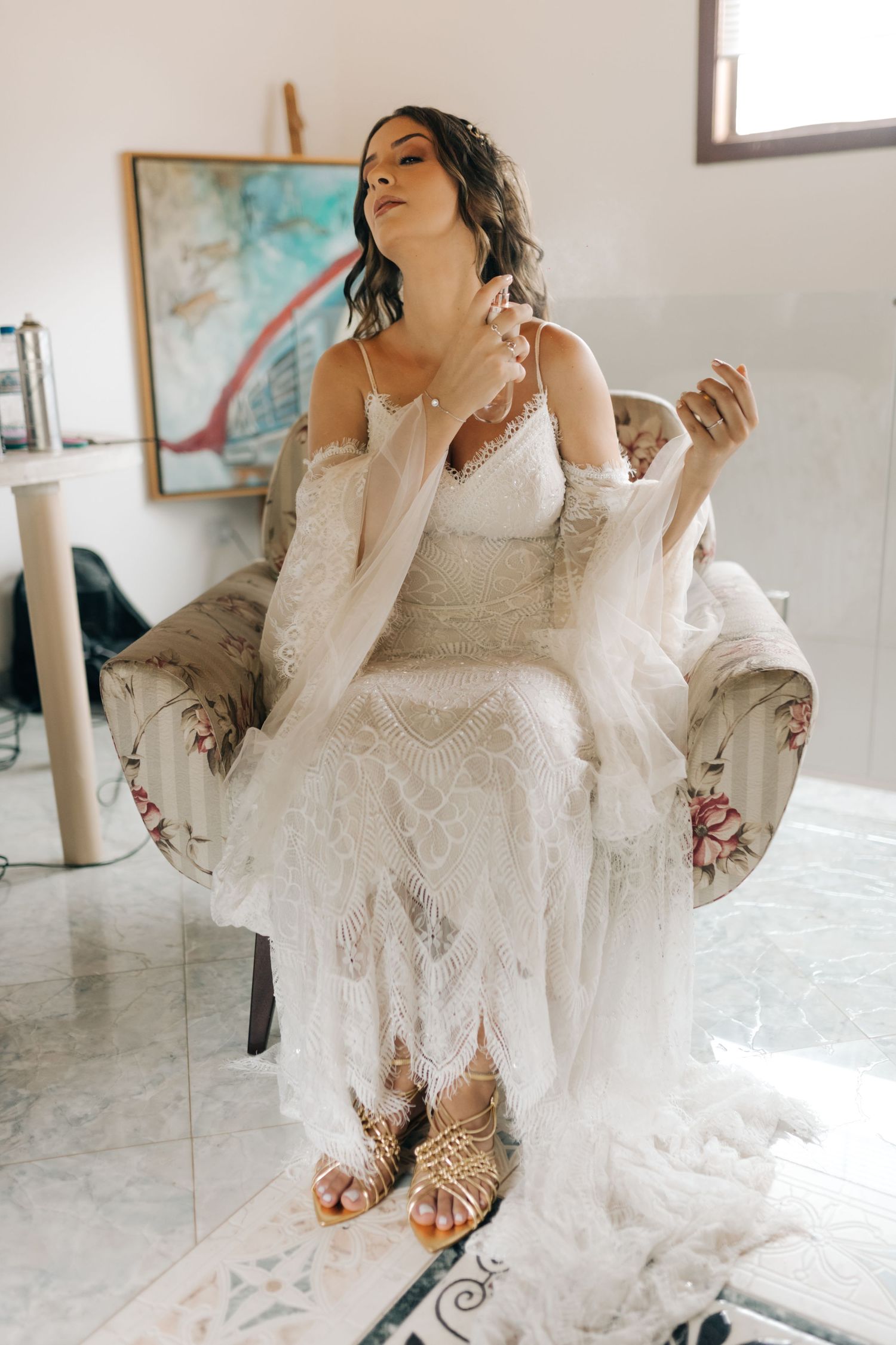Bride seated in floral armchair wearing lace dress and strappy sandals.