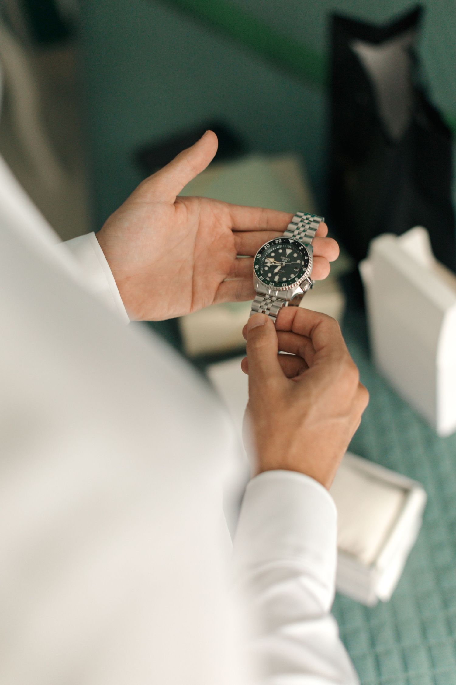 Close-up of hands holding a silver metal watch with dark face against white shirt.