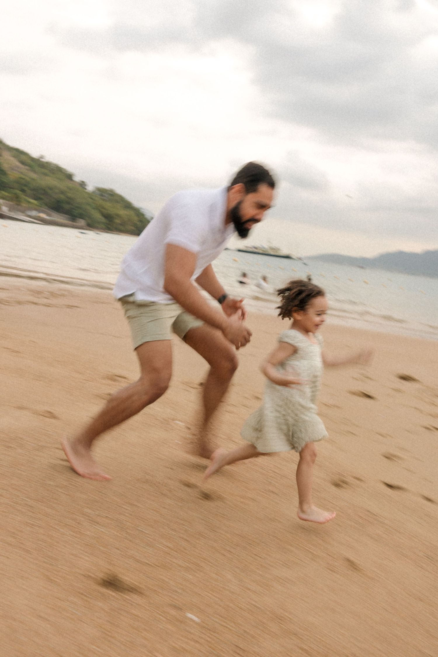 Two people run joyfully along a sandy beach with mountains visible in the background.
