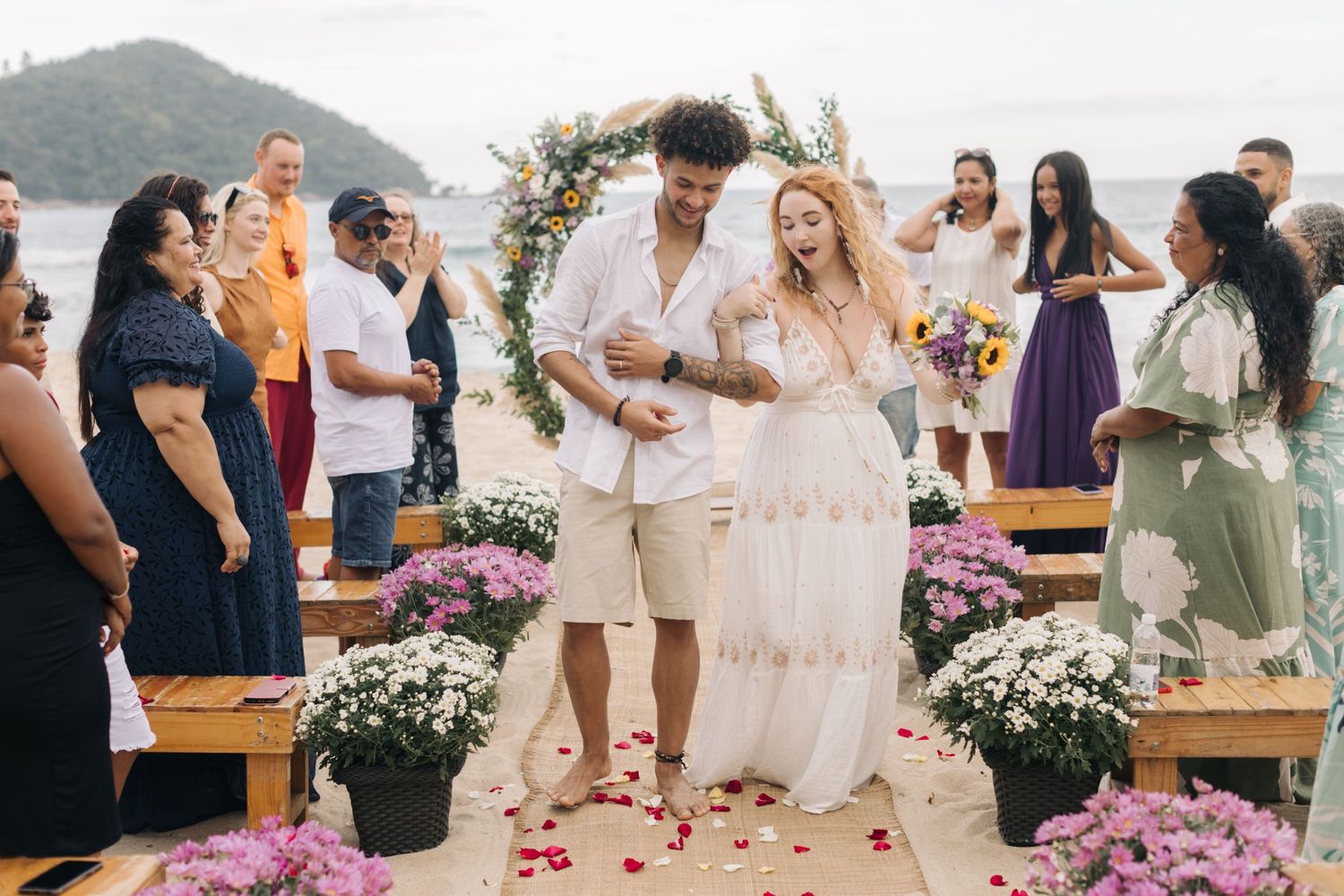 A joyous beach wedding ceremony with guests watching as a couple celebrates among pink flowers and wooden benches.