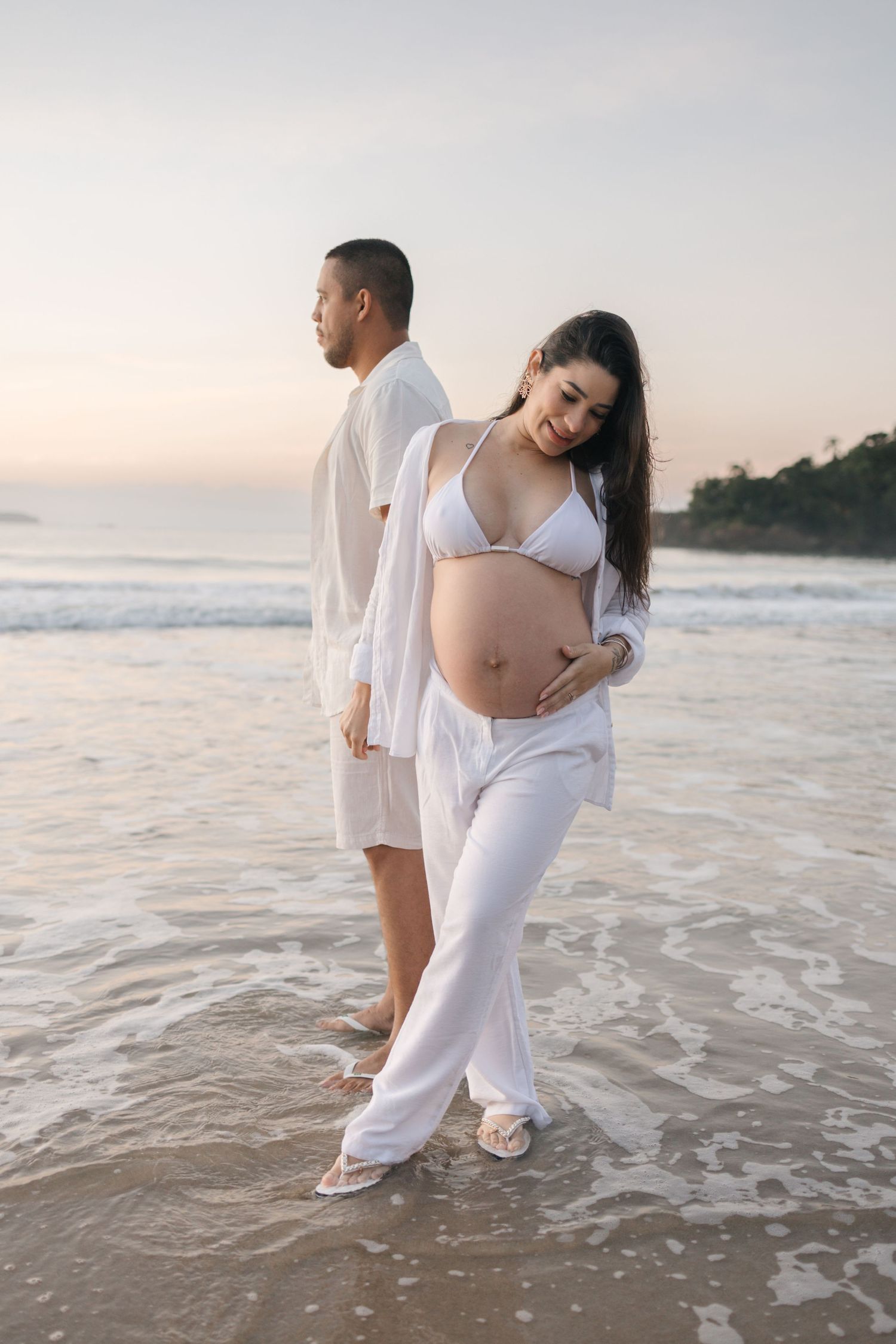 Couple in white clothing standing at ocean's edge during sunset maternity photoshoot.