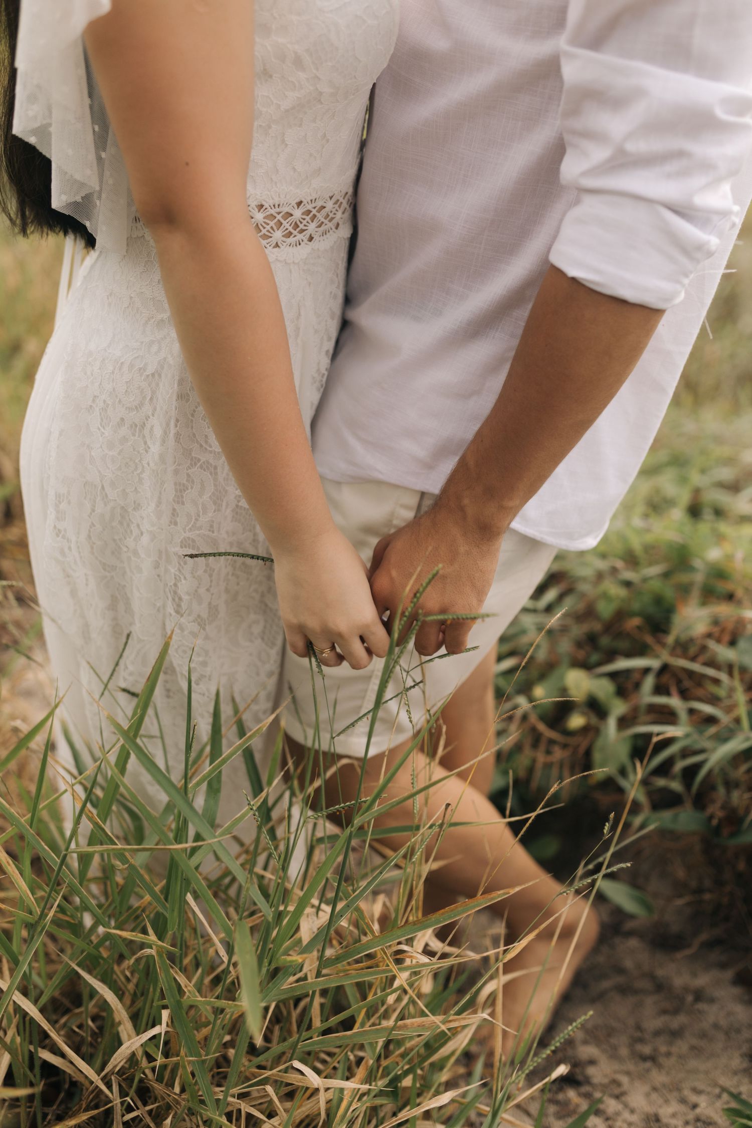 Close-up shot of couple's hands touching while standing in tall grass wearing white formal attire.