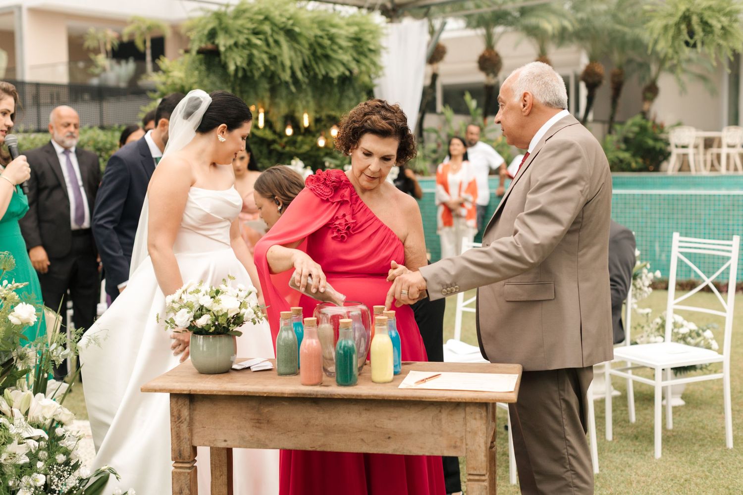 Wedding guests participate in a unity ceremony around a rustic wooden table with colored bottles.