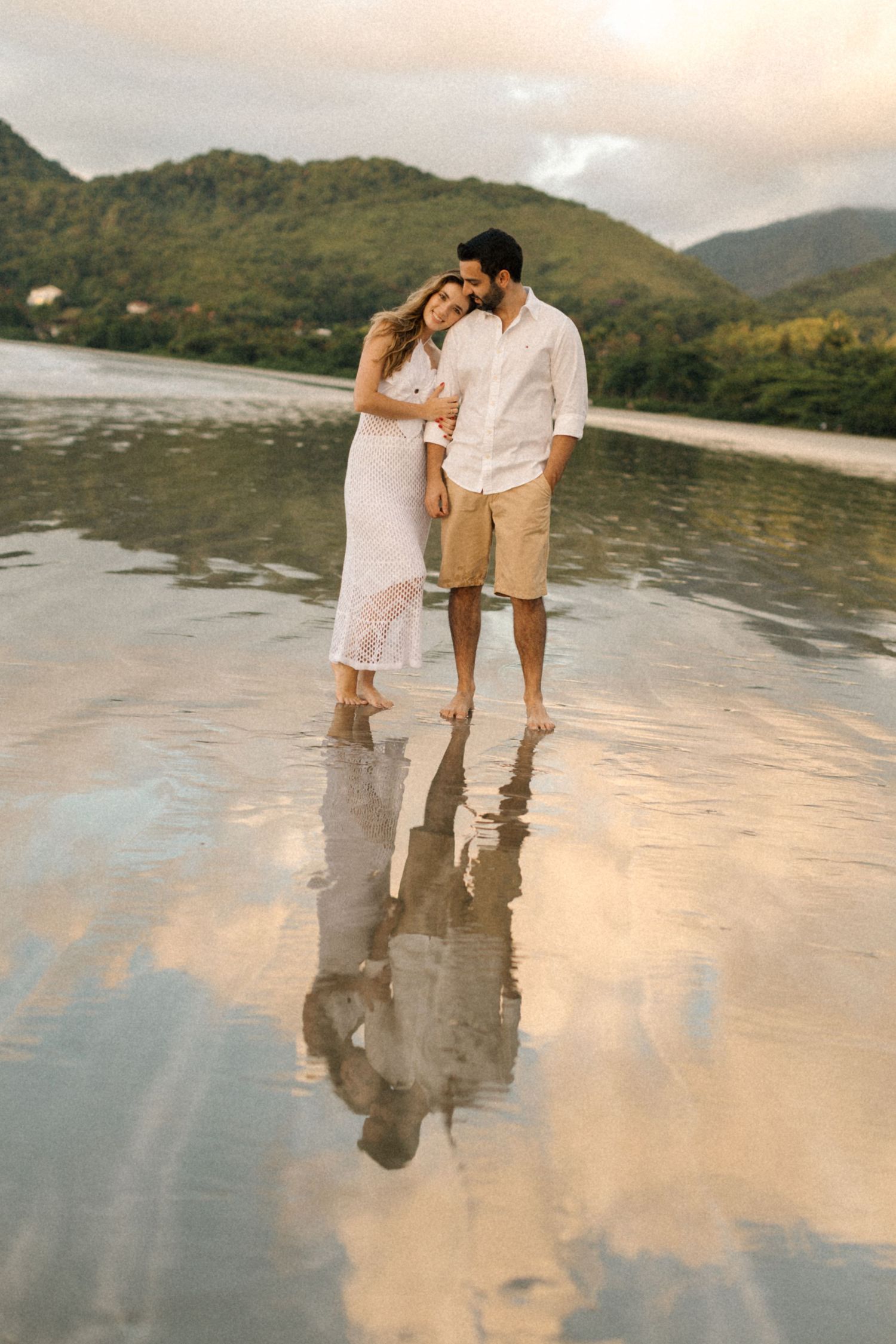 Couple walking hand in hand on reflective wet beach with mountains in background at sunset.