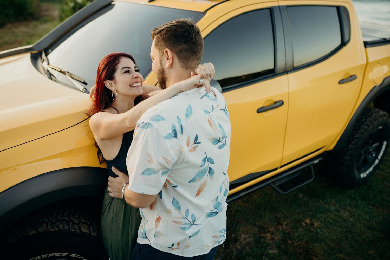 Couple embracing by yellow pickup truck with coastal scenery in background.