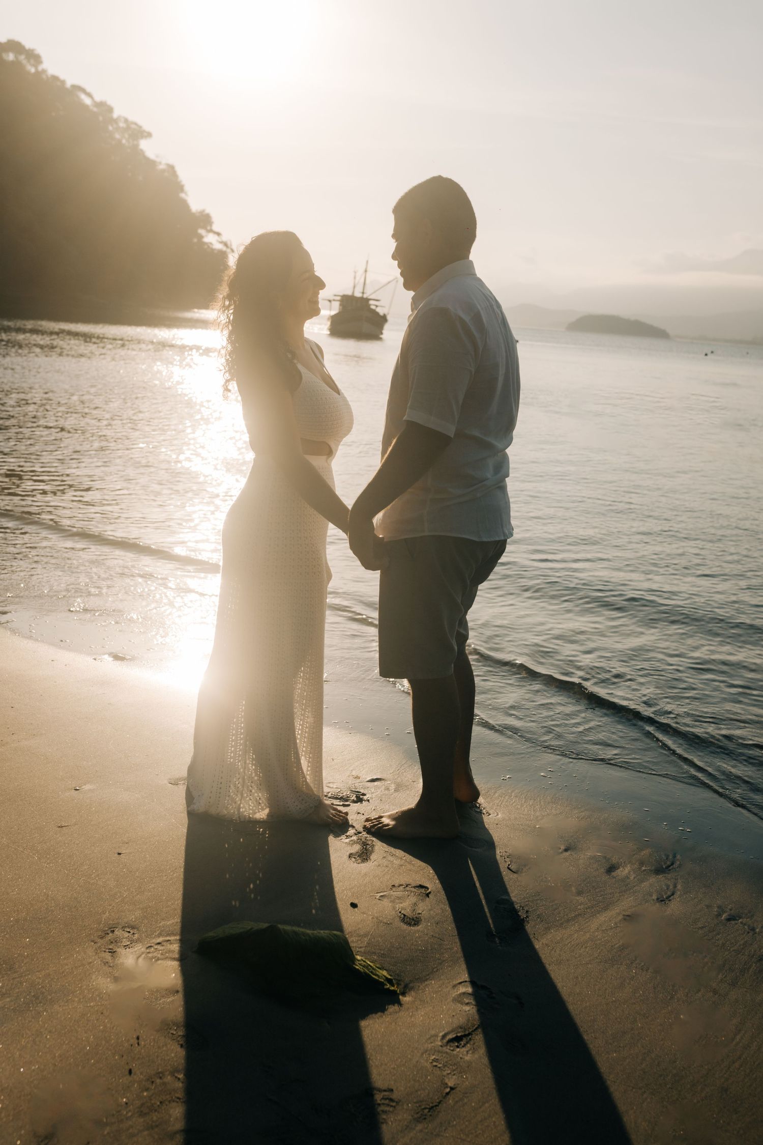 Couple standing on beach during golden hour with boats and mountains in background.