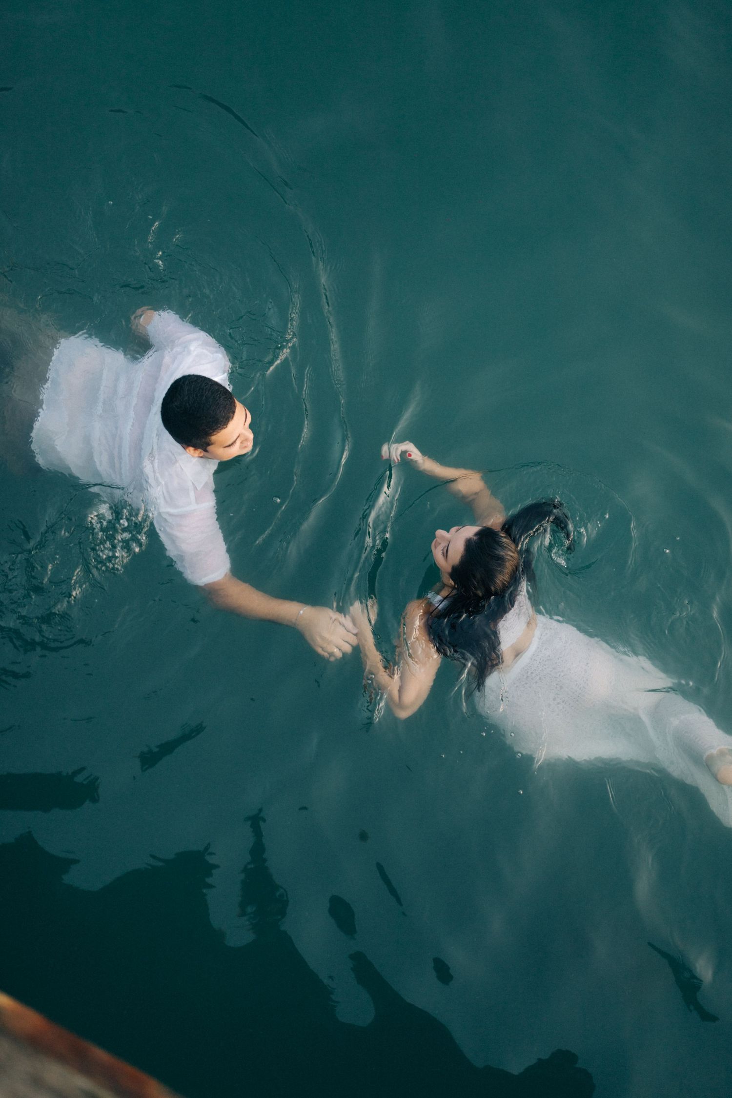 A couple floats in turquoise water while holding hands, viewed from above.