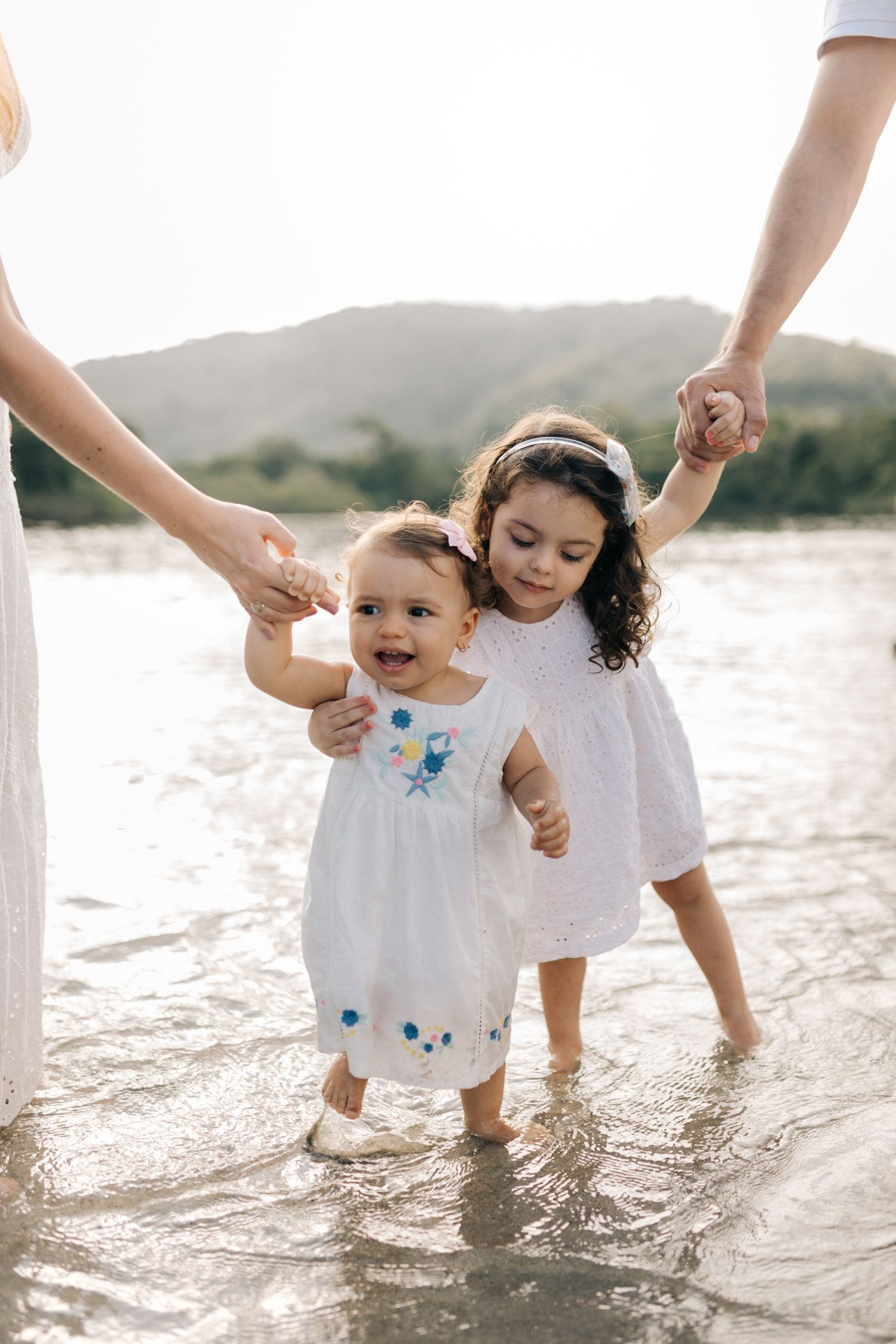 Small children playing in shallow water while holding adults' hands at the beach.