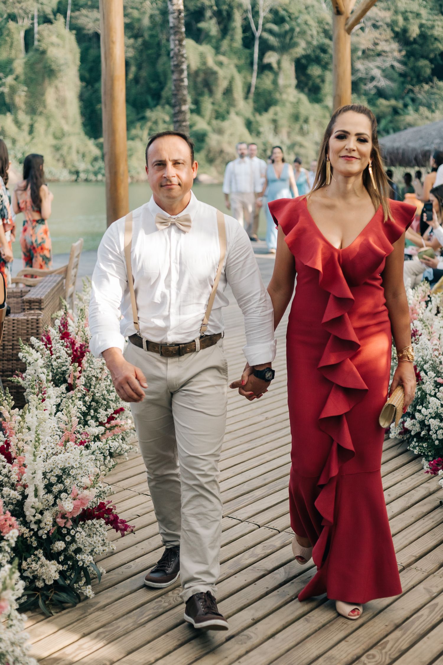 Elegantly dressed couple walking on wooden boardwalk decorated with floral arrangements by water.