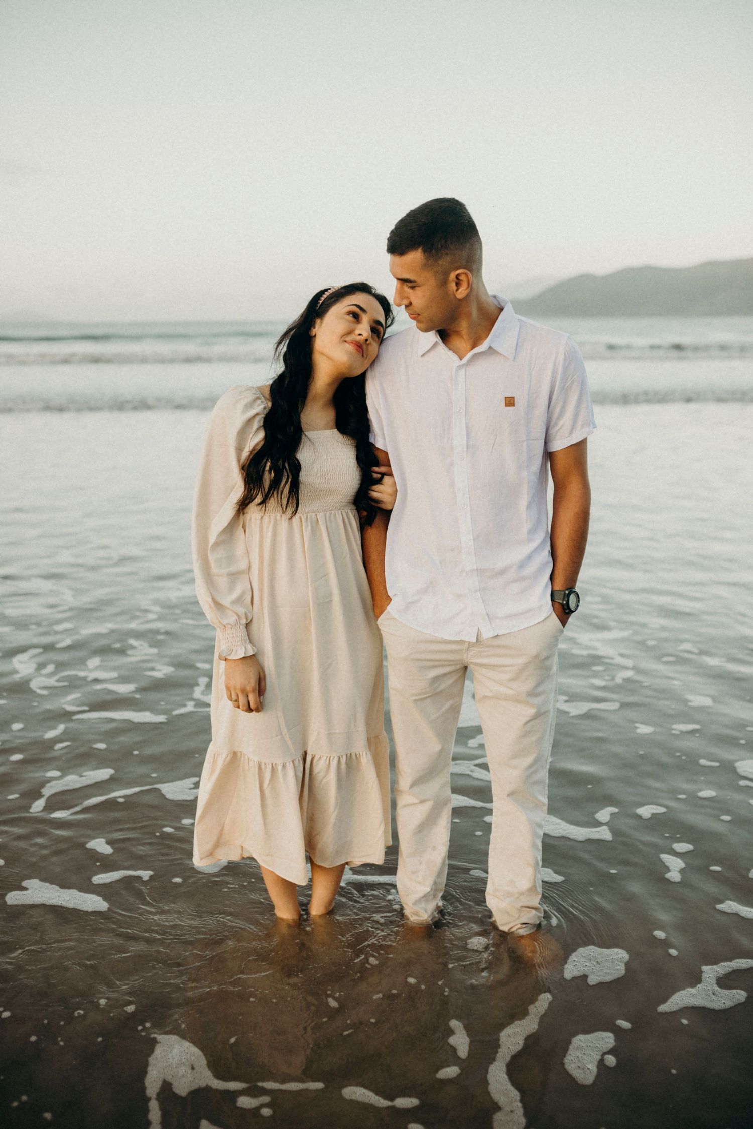 A couple in matching light outfits shares an intimate moment while standing in shallow beach water.