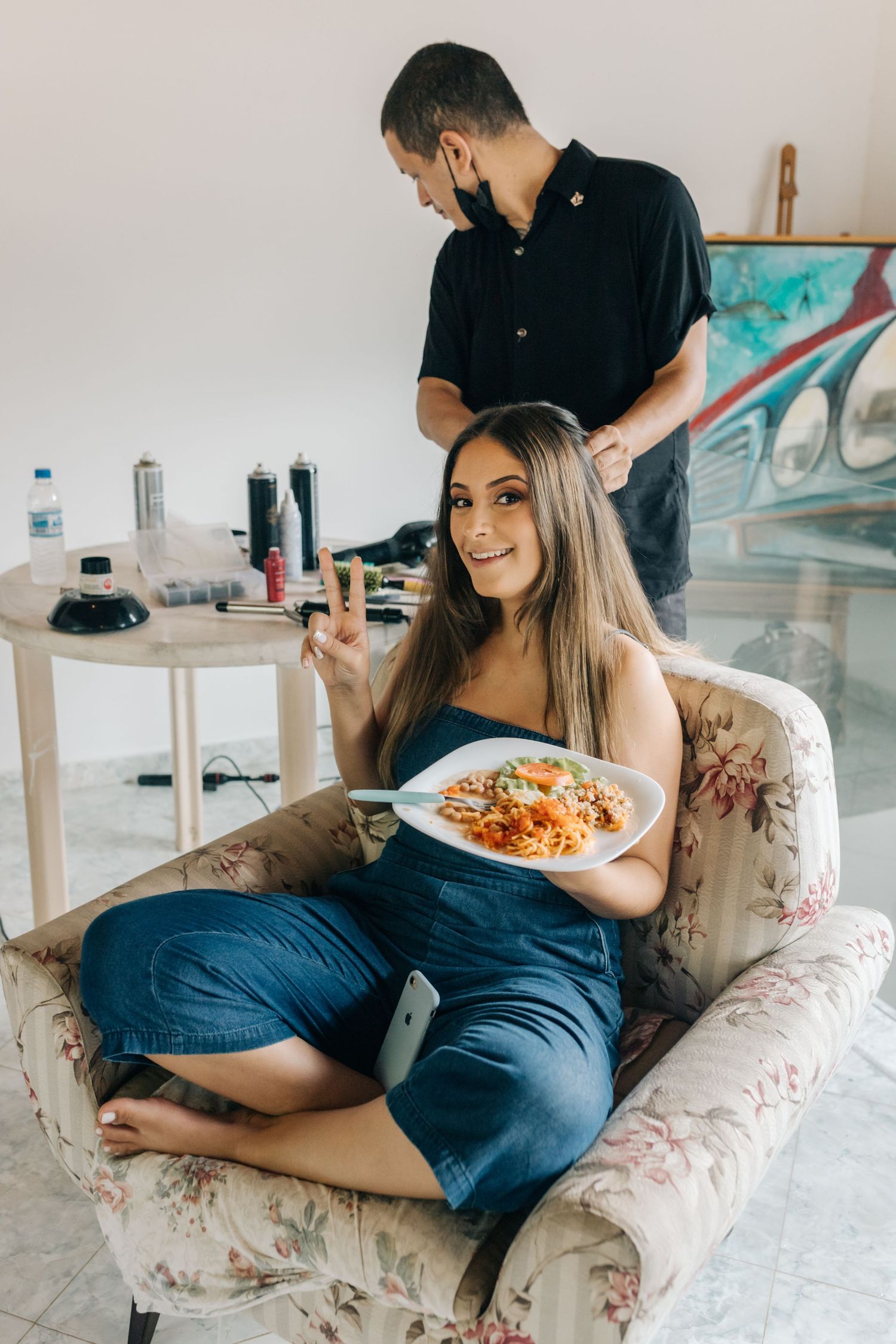Stylist working on client's hair while she enjoys pasta in a floral armchair.