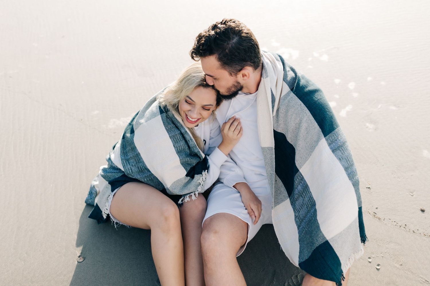 A couple wrapped in a blanket share an intimate moment while sitting together on a beach.