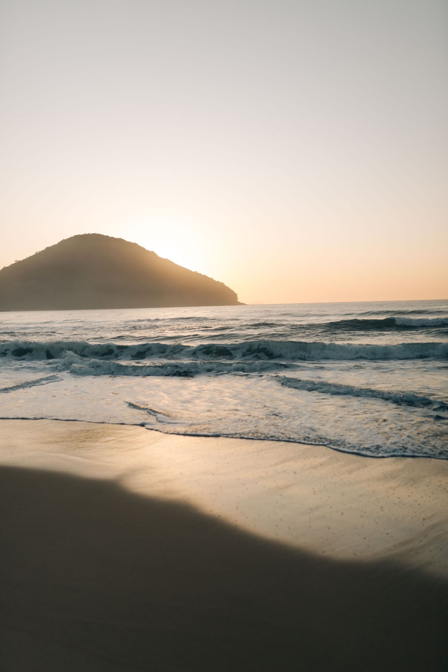 Serene sunset beach landscape with rolling waves and mountain silhouette against a pastel sky.