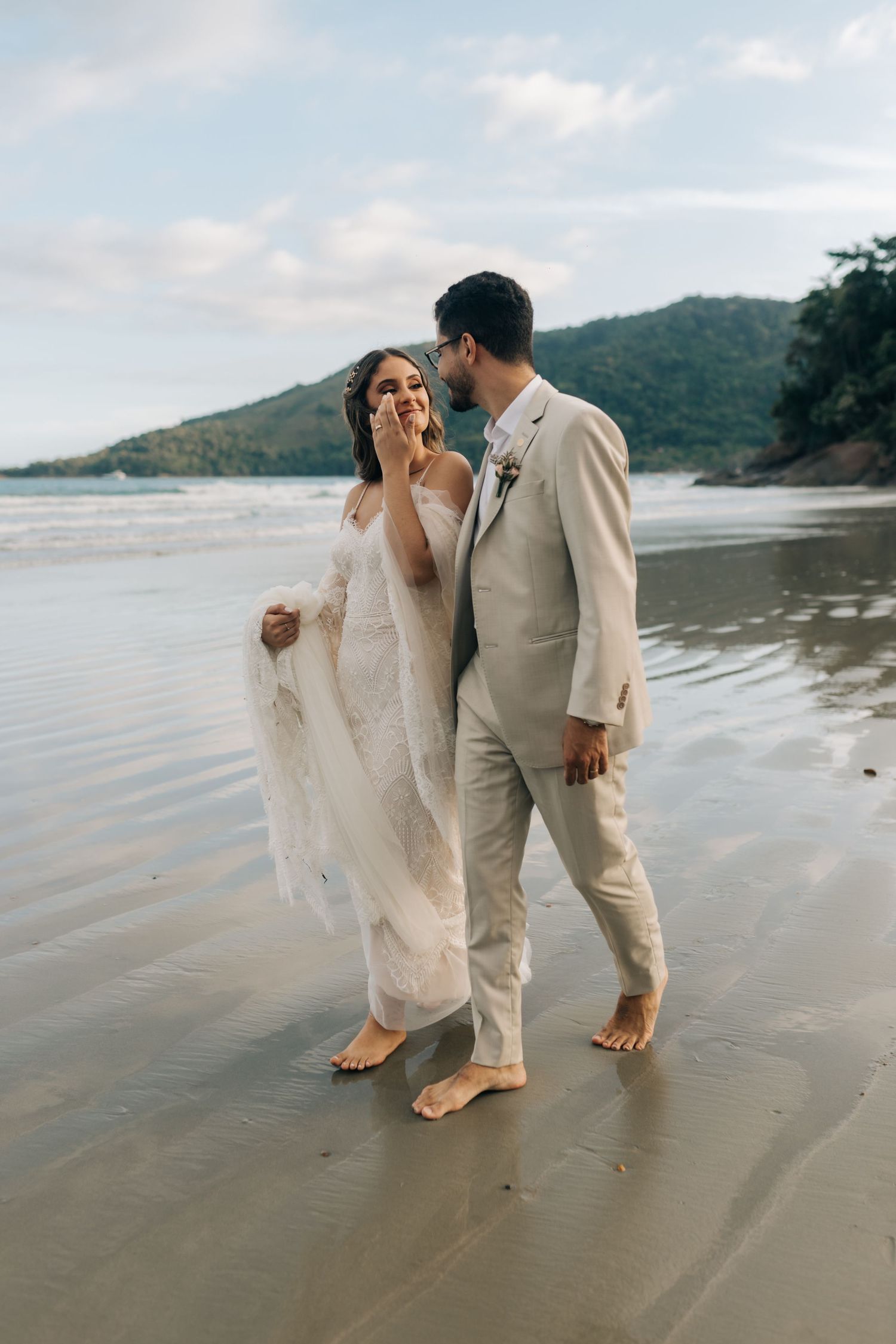 Candid beach wedding moment of couple laughing while walking barefoot along the shoreline.