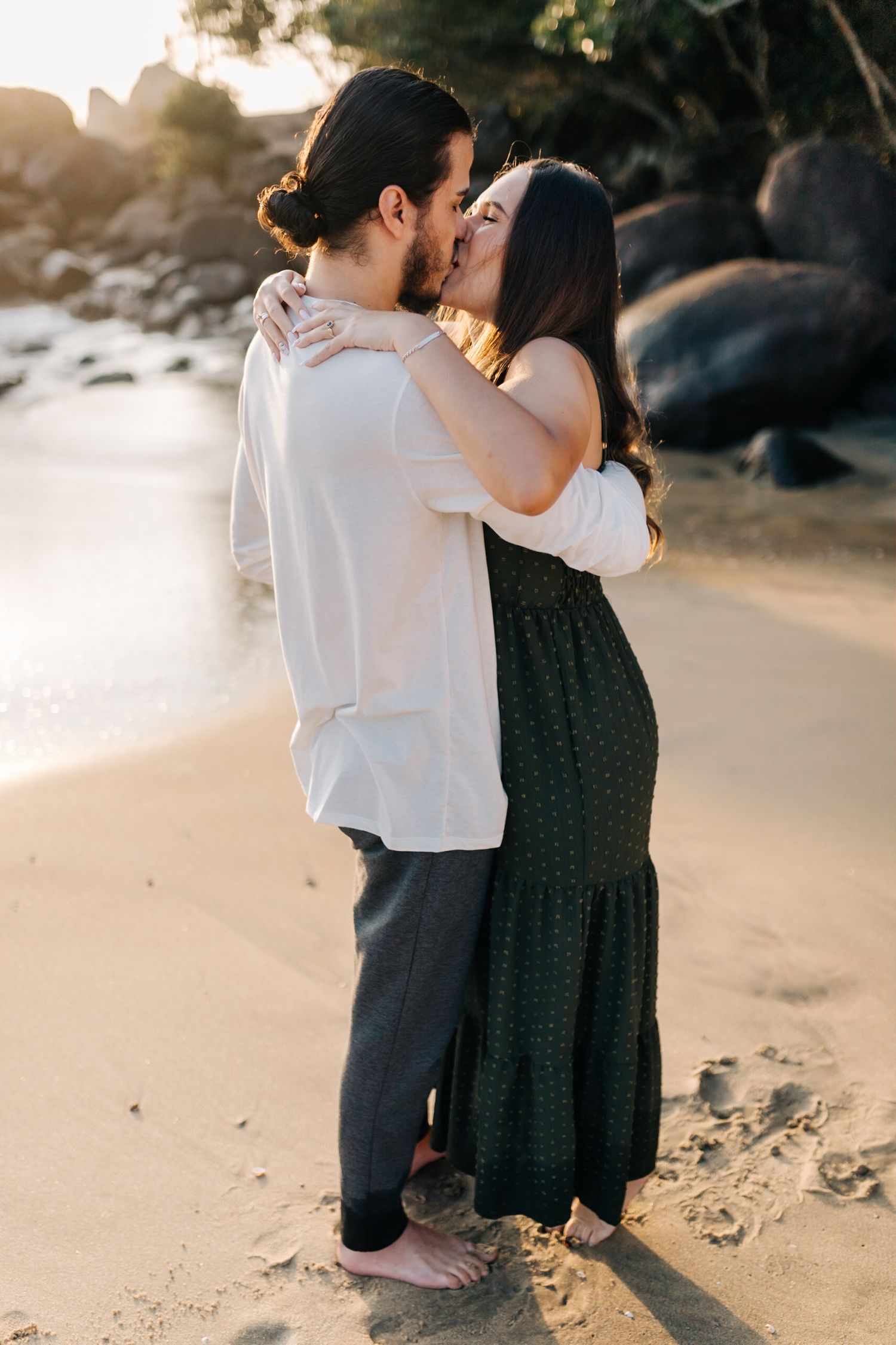 Romantic couple embracing on a sunlit beach with rocky coastline in the background.