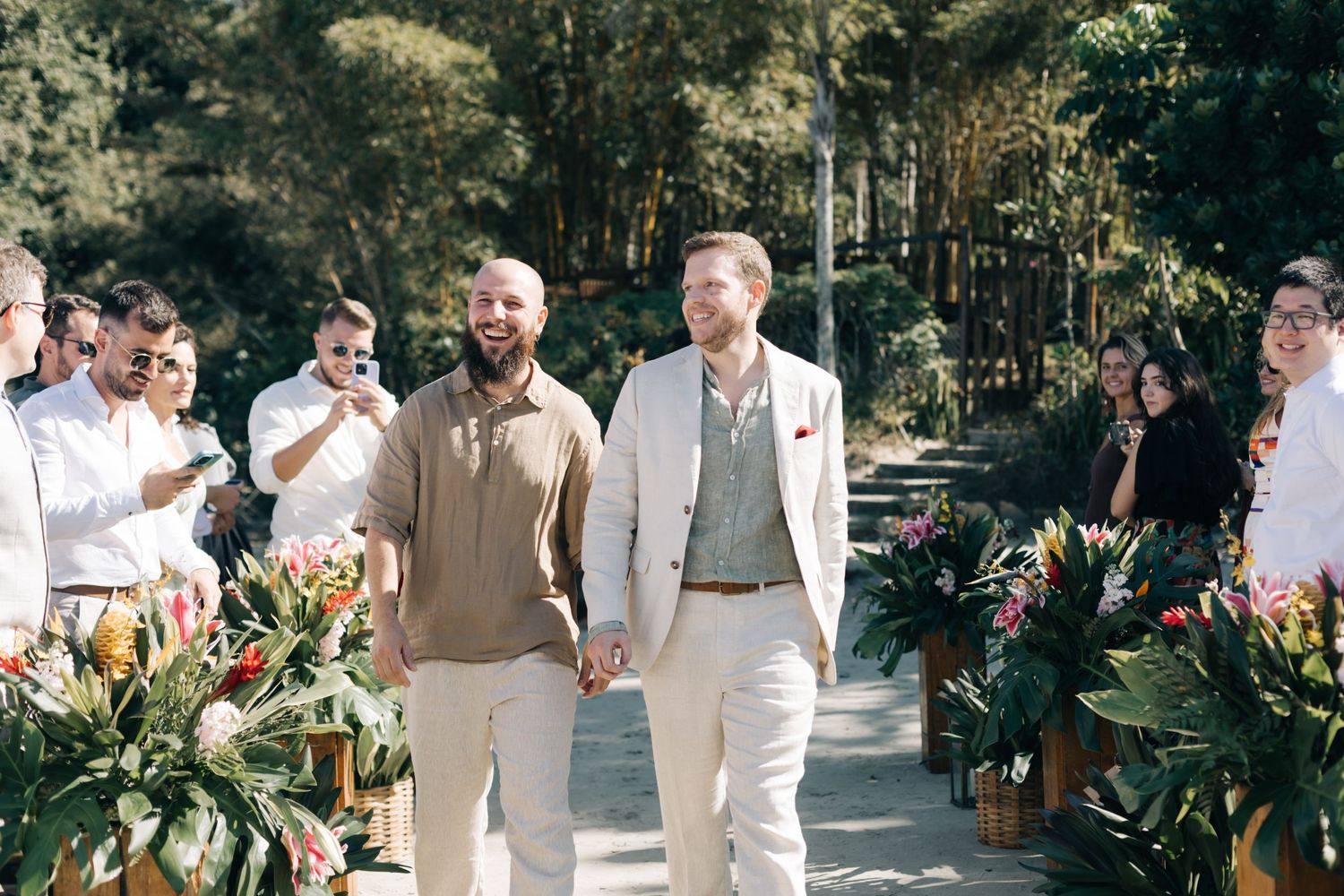 Wedding party members in neutral tones gather on a sunlit garden pathway lined with tropical plants.