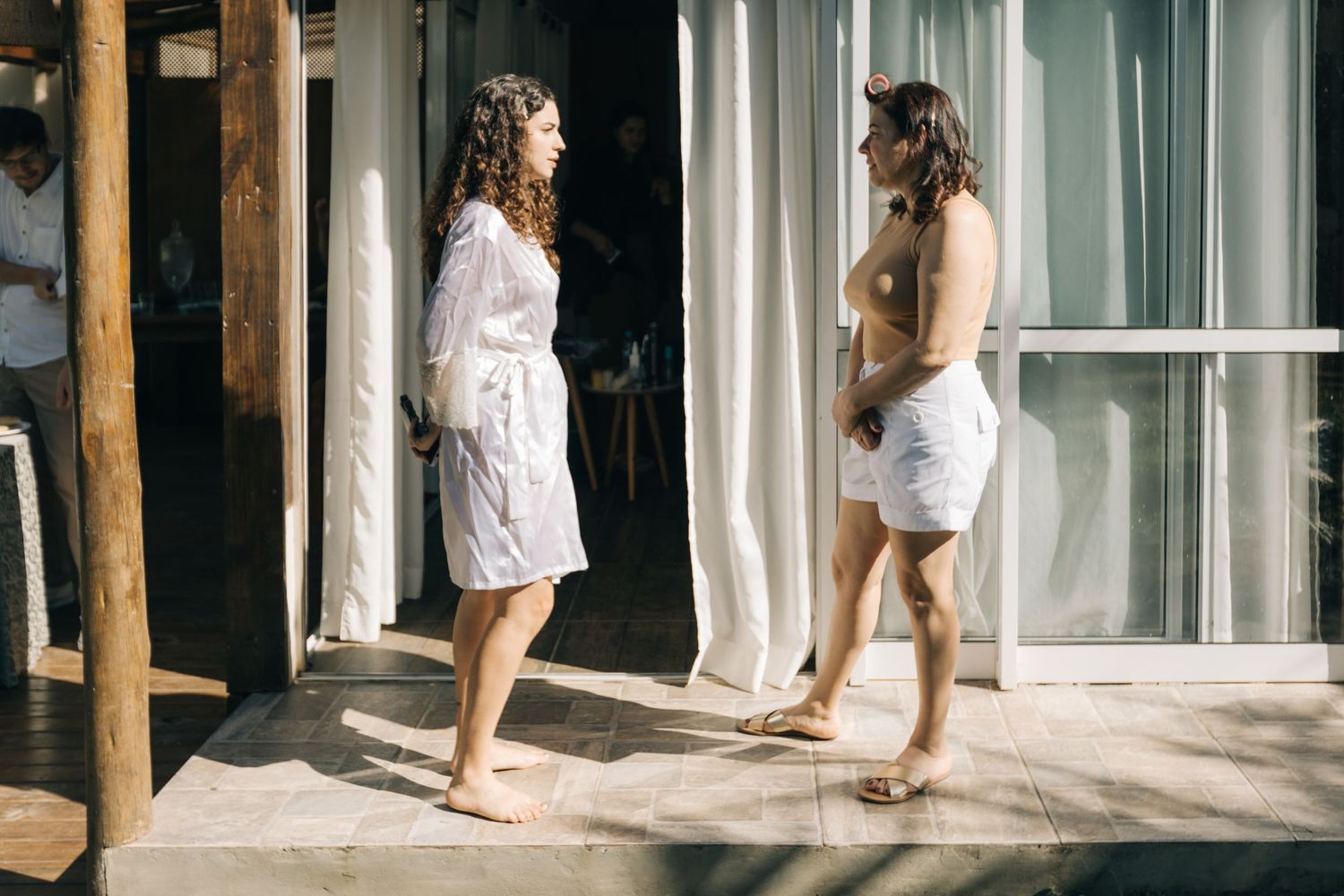 Two figures in white robes chatting on sunny porch with sheer curtains.