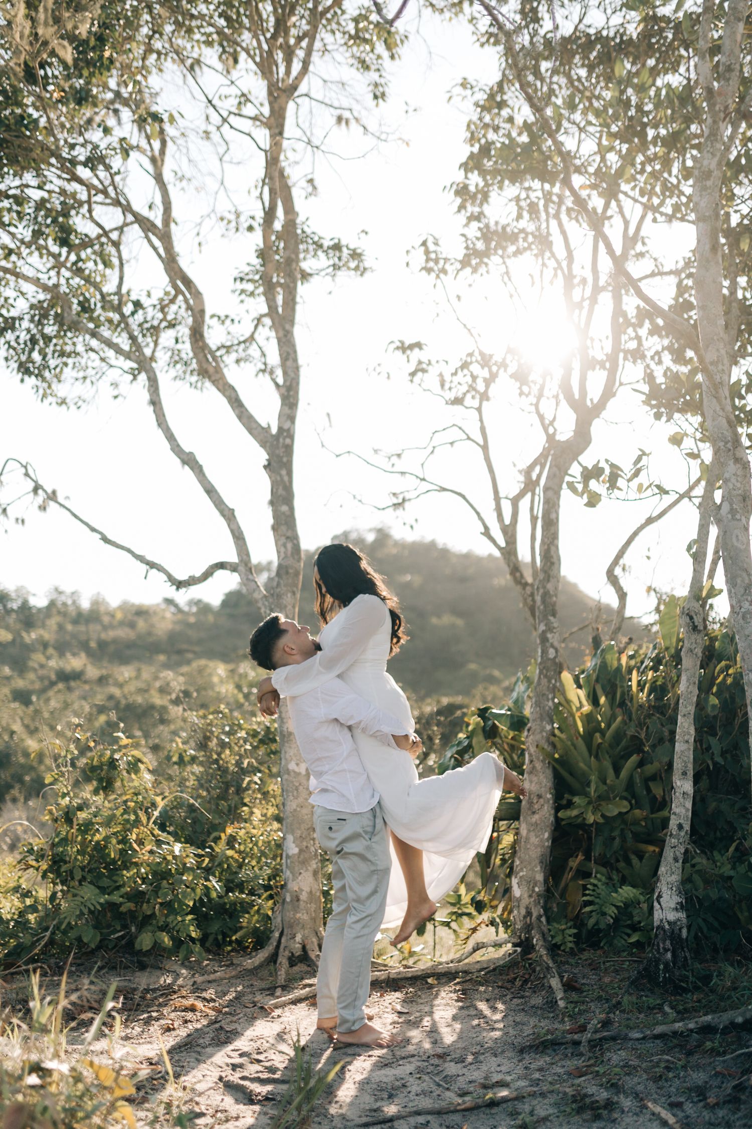 A romantic couple in white outfits embrace against sunlit trees in a dreamy outdoor setting.