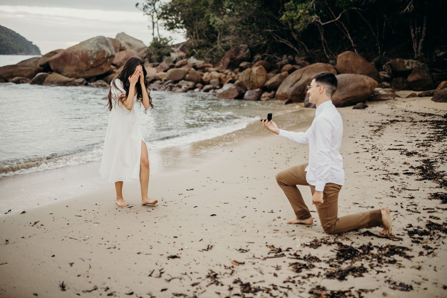 Marriage proposal moment captured on sandy beach with ocean views.