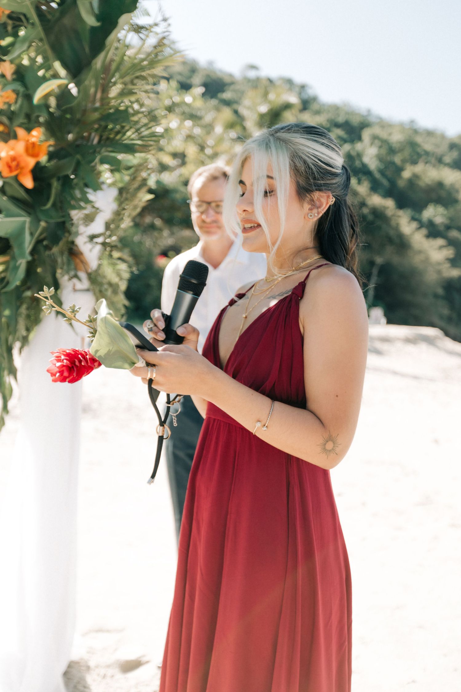 Wedding guest in burgundy dress giving speech at outdoor ceremony.