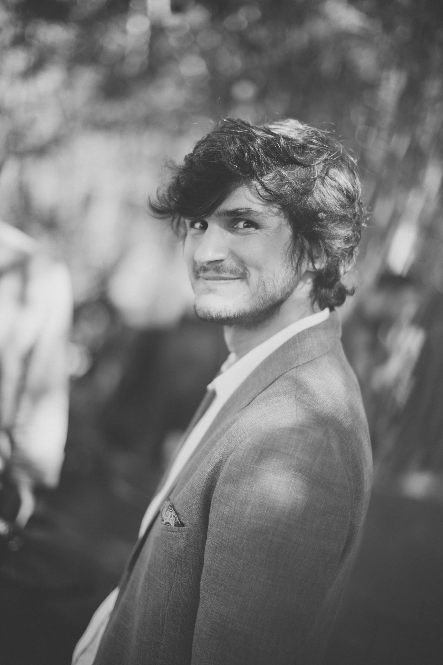 Black and white portrait of person in suit smiling against blurred natural background.