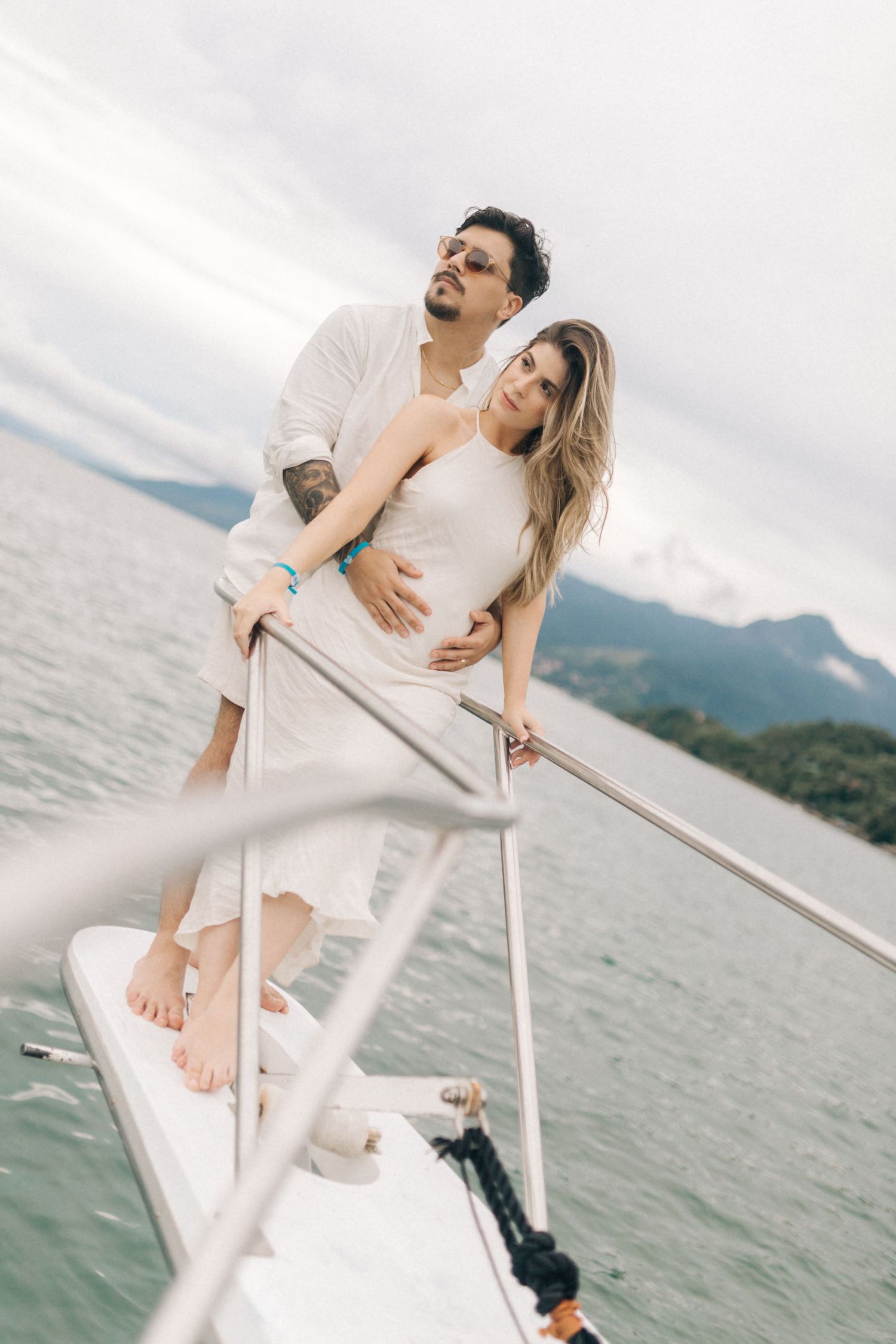 Couple embracing on yacht bow with mountainous coastline in background.
