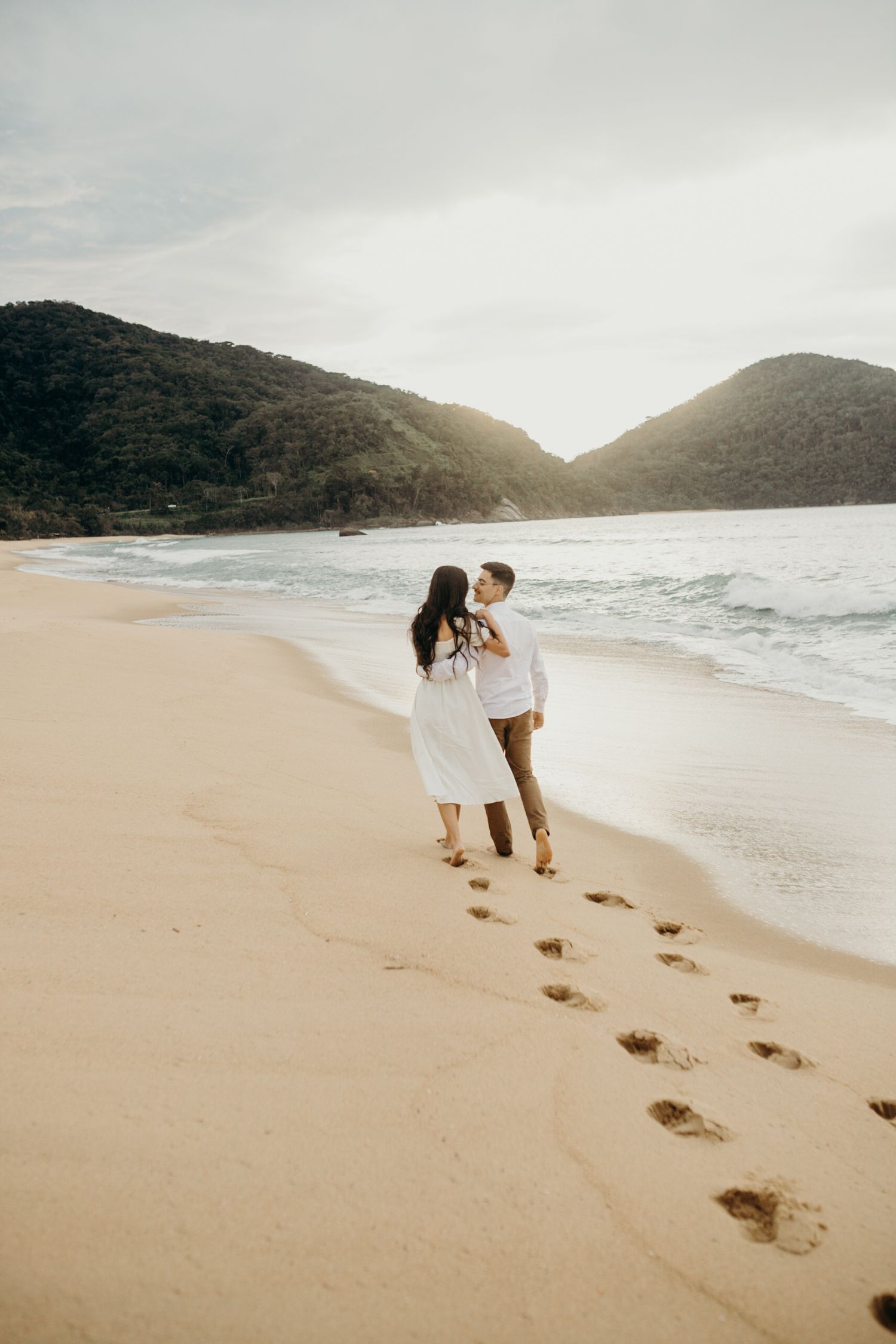 Footprints trail behind a couple walking along a pristine beach with mountains and ocean views.