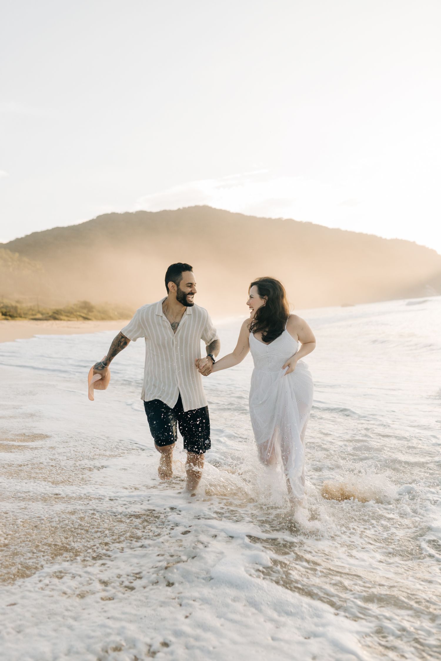 Couple splashing and playing in ocean waves during a beach photoshoot at sunset.