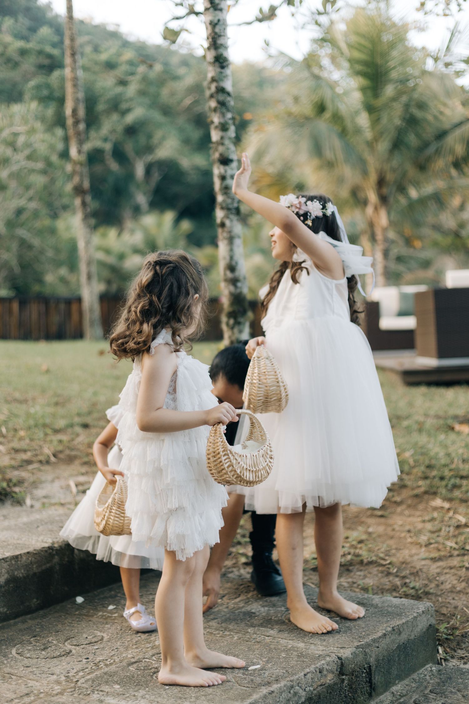 Two young attendants in white dresses carry woven baskets at outdoor event.