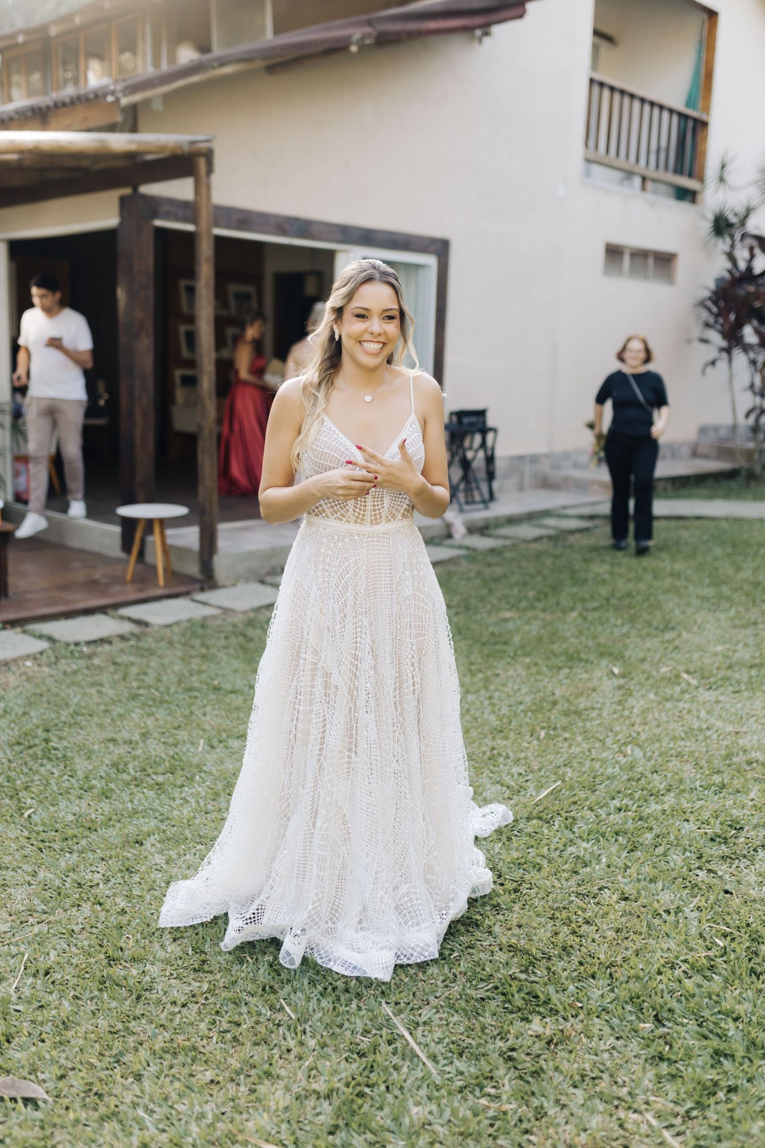 A bride wearing a delicate white lace wedding gown stands on grass outside a building.