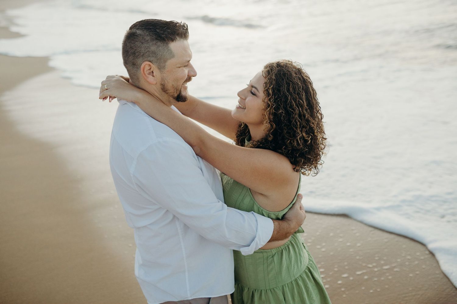 A couple embraces romantically on a sandy beach at sunset wearing white and green outfits.